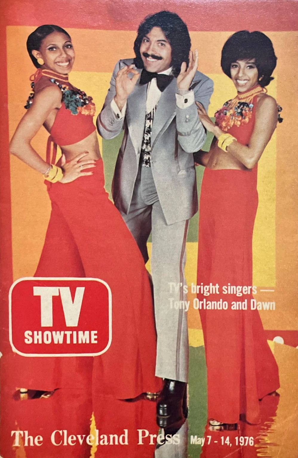 A retro magazine cover featuring Tony Orlando and the duo Dawn, showcasing them in colorful outfits. Tony, dressed in a grey suit with a bow tie, poses playfully between two women in red dresses, both adorned with vibrant accessories, set against a bright, multicolored background.