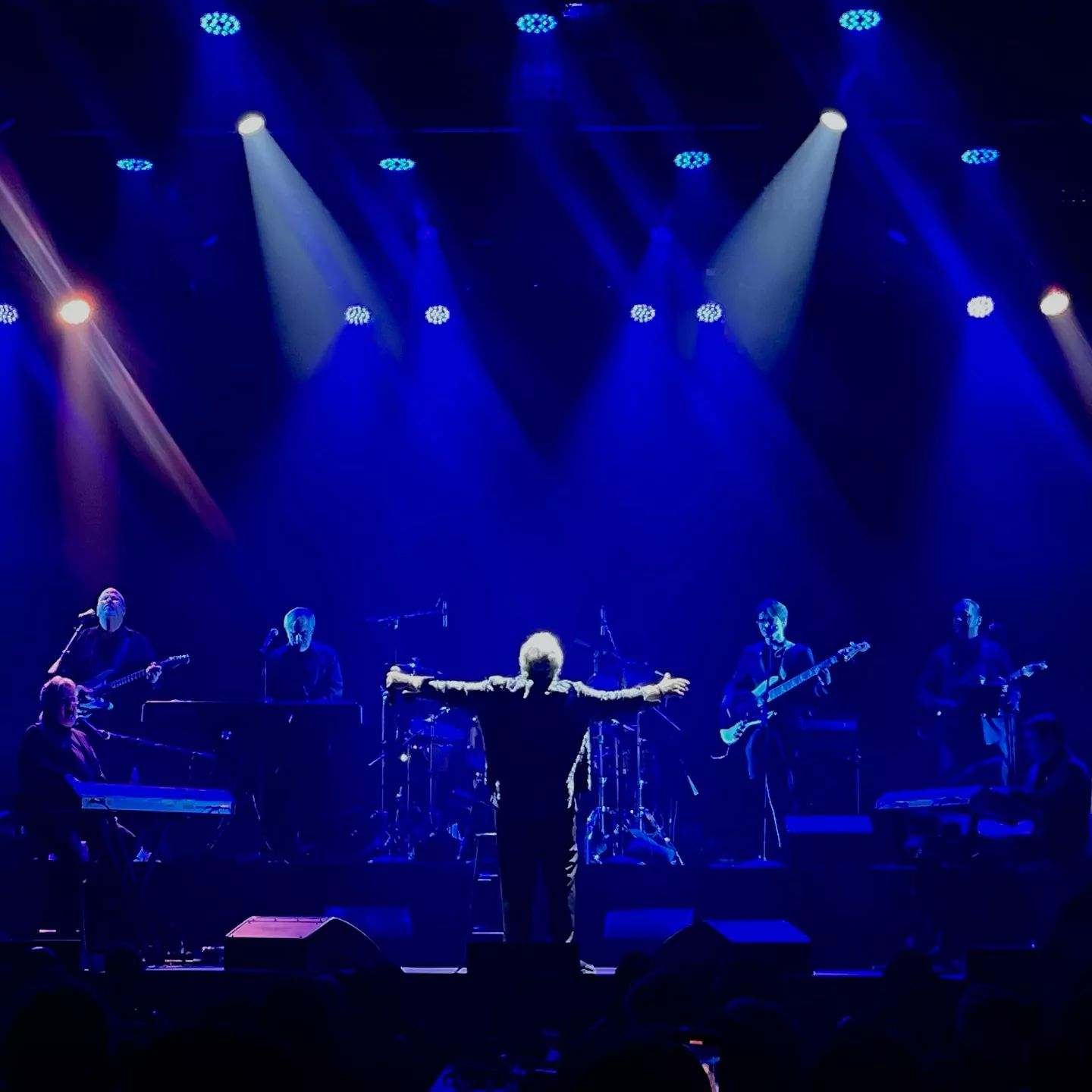 A musician performing on stage with arms outstretched, surrounded by a band in blue lighting.