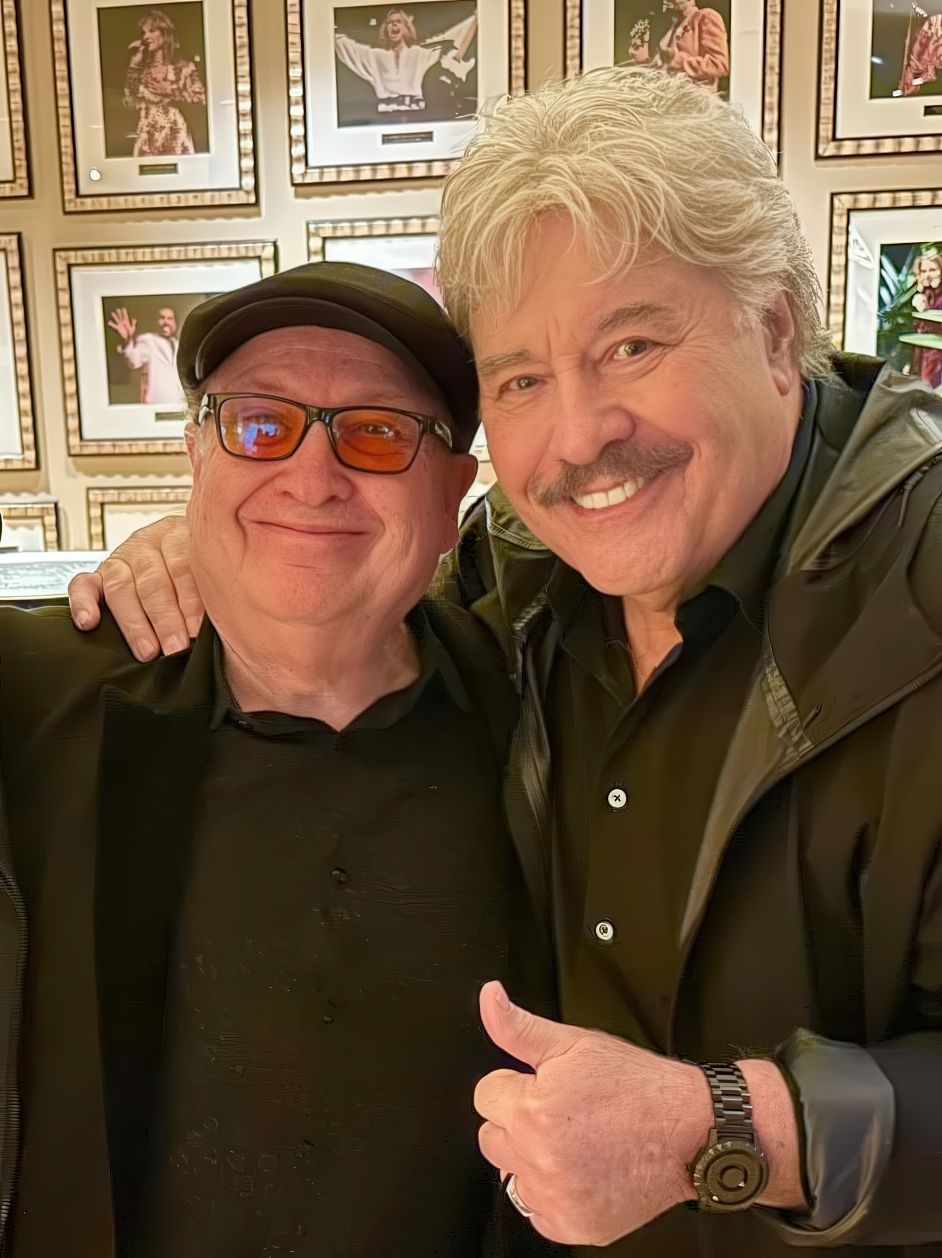 Tony Orlando and a friend posing together, smiling, with framed photographs of performers in the background.