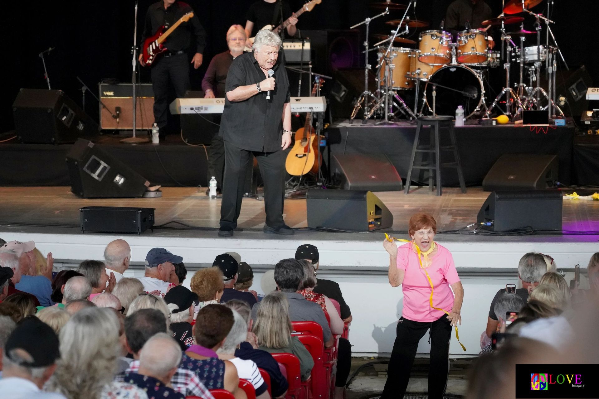 A lively performance scene featuring an older man singing on stage with a microphone, while an enthusiastic audience reacts. A woman in a pink shirt is dancing in the foreground, wearing a yellow ribbon around her neck, creating a joyful atmosphere.