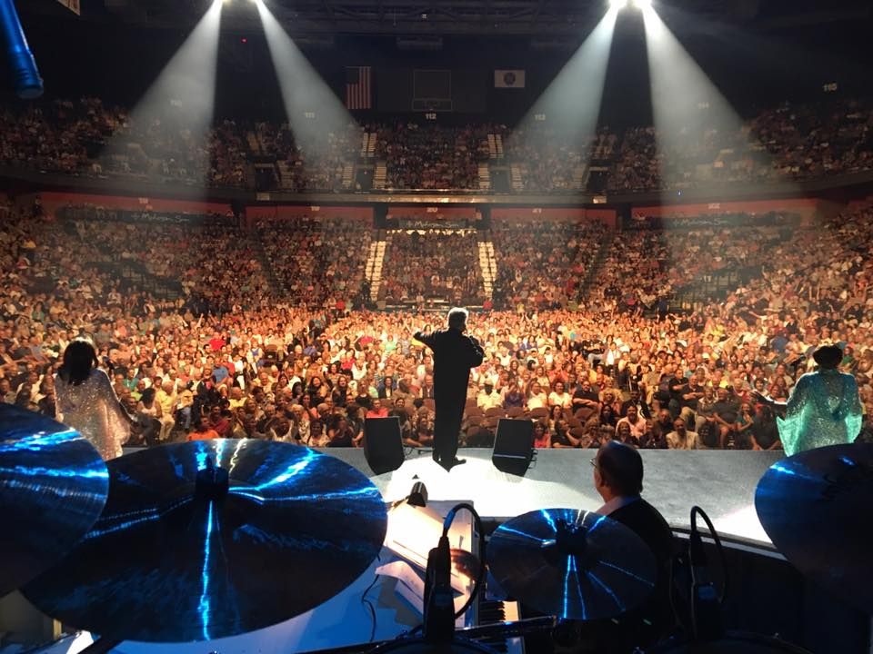 View of a large audience cheering at a concert, with a performer on stage and band members in the foreground, illuminated by stage lights.