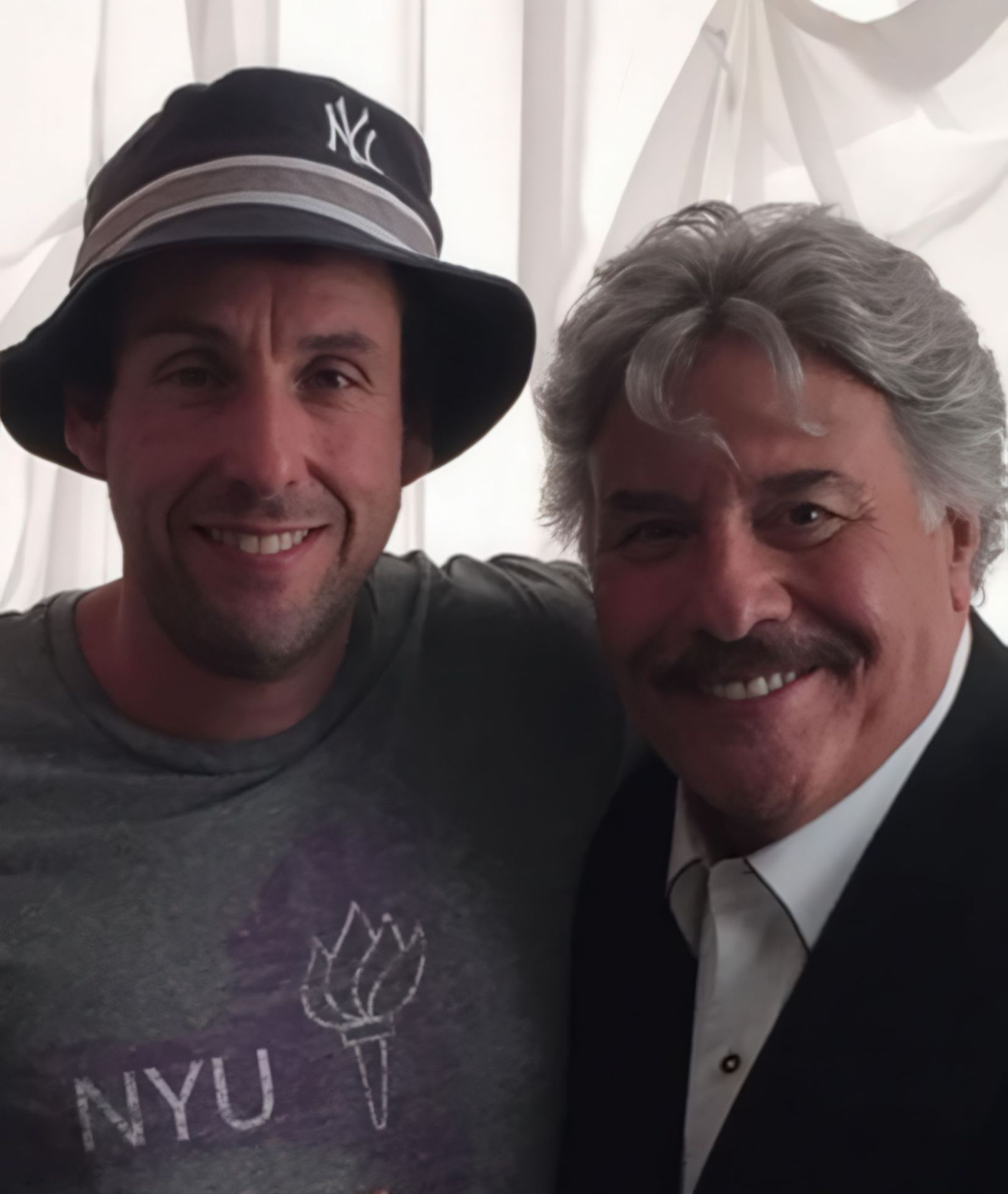 A smiling man wearing a grey NYU t-shirt and a bucket hat stands next to another man in a white shirt and black suit, both posing for a photo against a light-colored background.