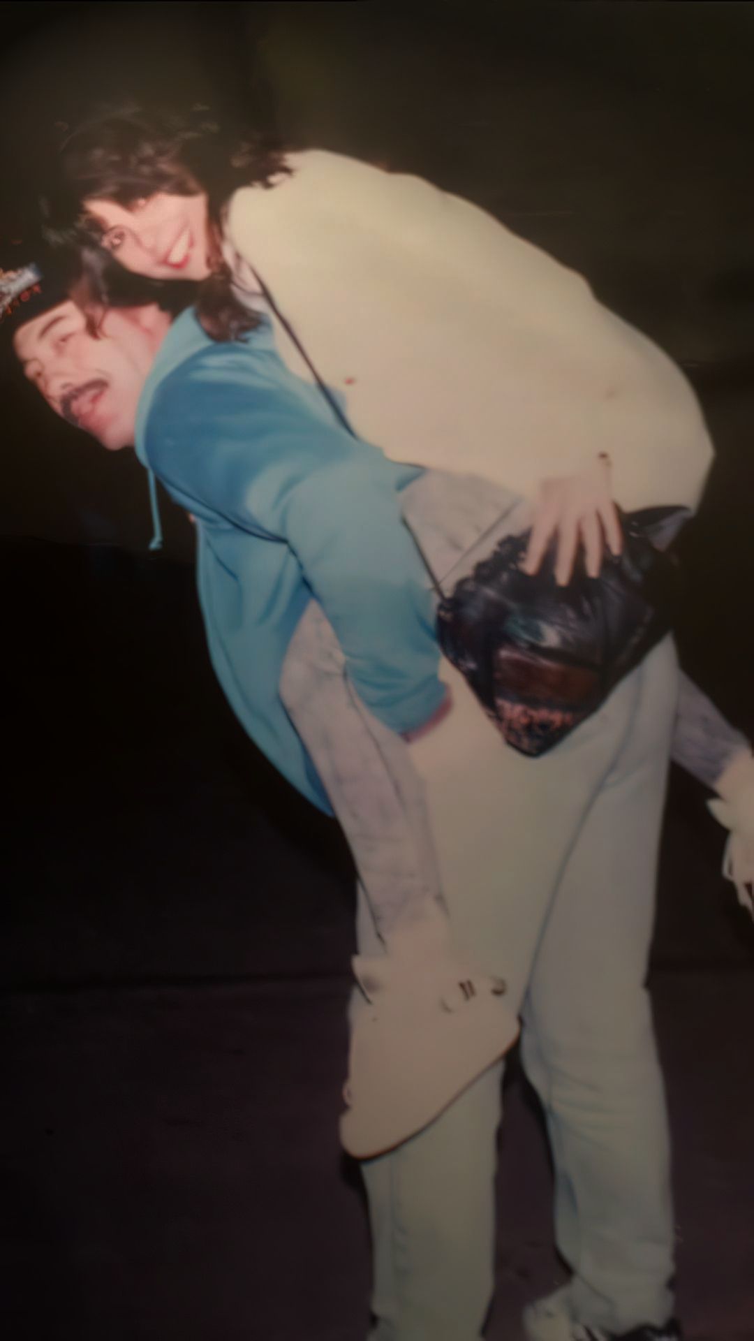 A man in a blue jacket is giving a piggyback ride to a smiling woman with long hair, who is wearing a light-colored top. The background is dimly lit.