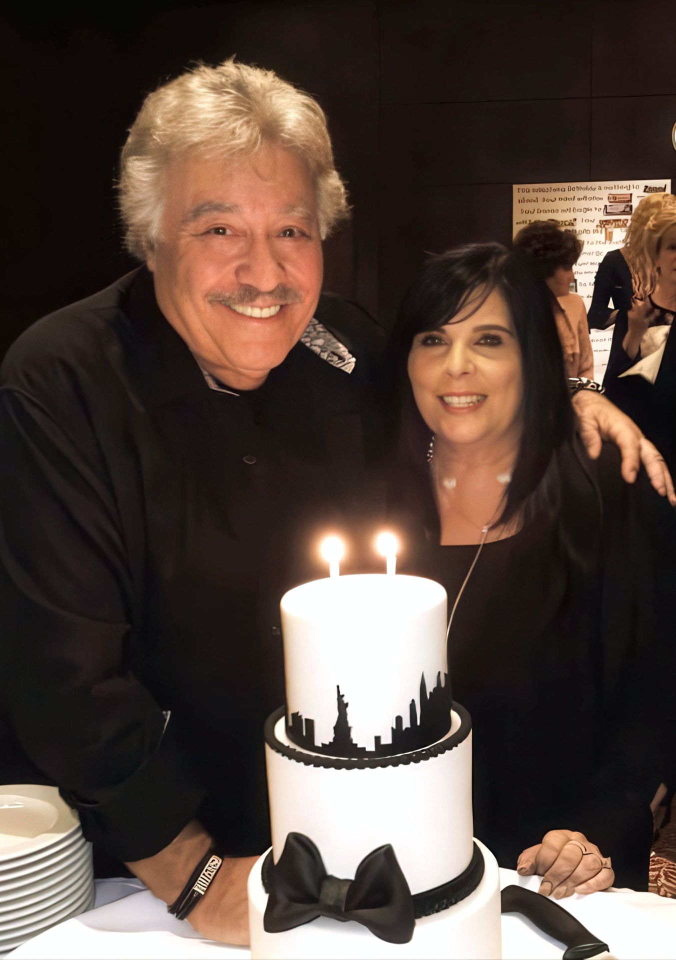 Tony Orlando and a woman pose together by a decorative cake with lit candles, celebrating a special occasion.