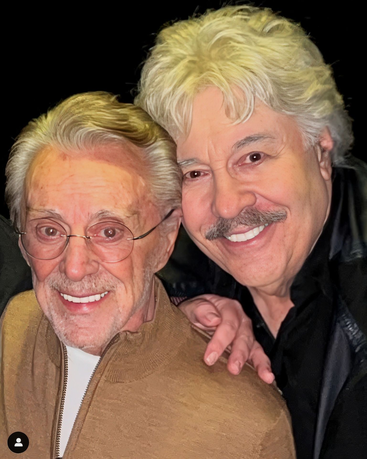 Two smiling men posing closely together, one with light-colored hair and a mustache, and the other with glasses and gray hair, both displaying warm expressions.