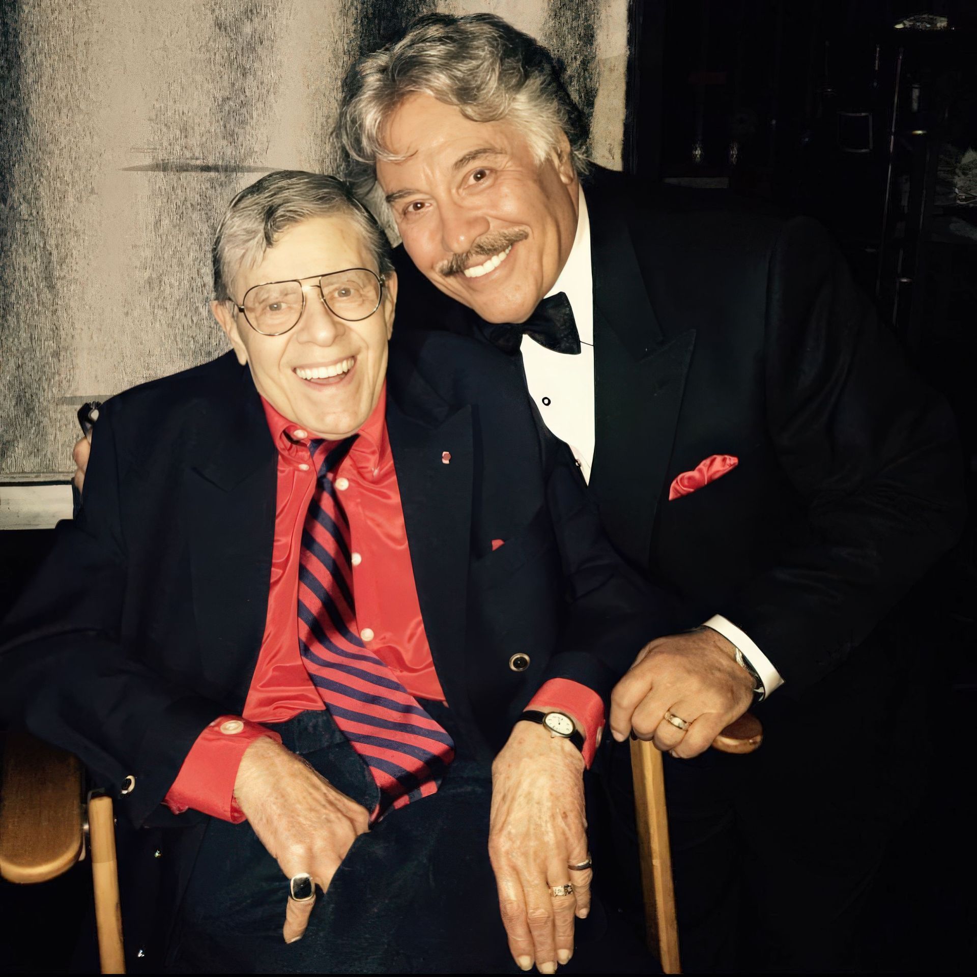 A smiling elderly man in a black blazer and red shirt with a striped tie sits in a chair, holding hands with another man in a tuxedo who stands beside him, both appearing joyful and engaged.