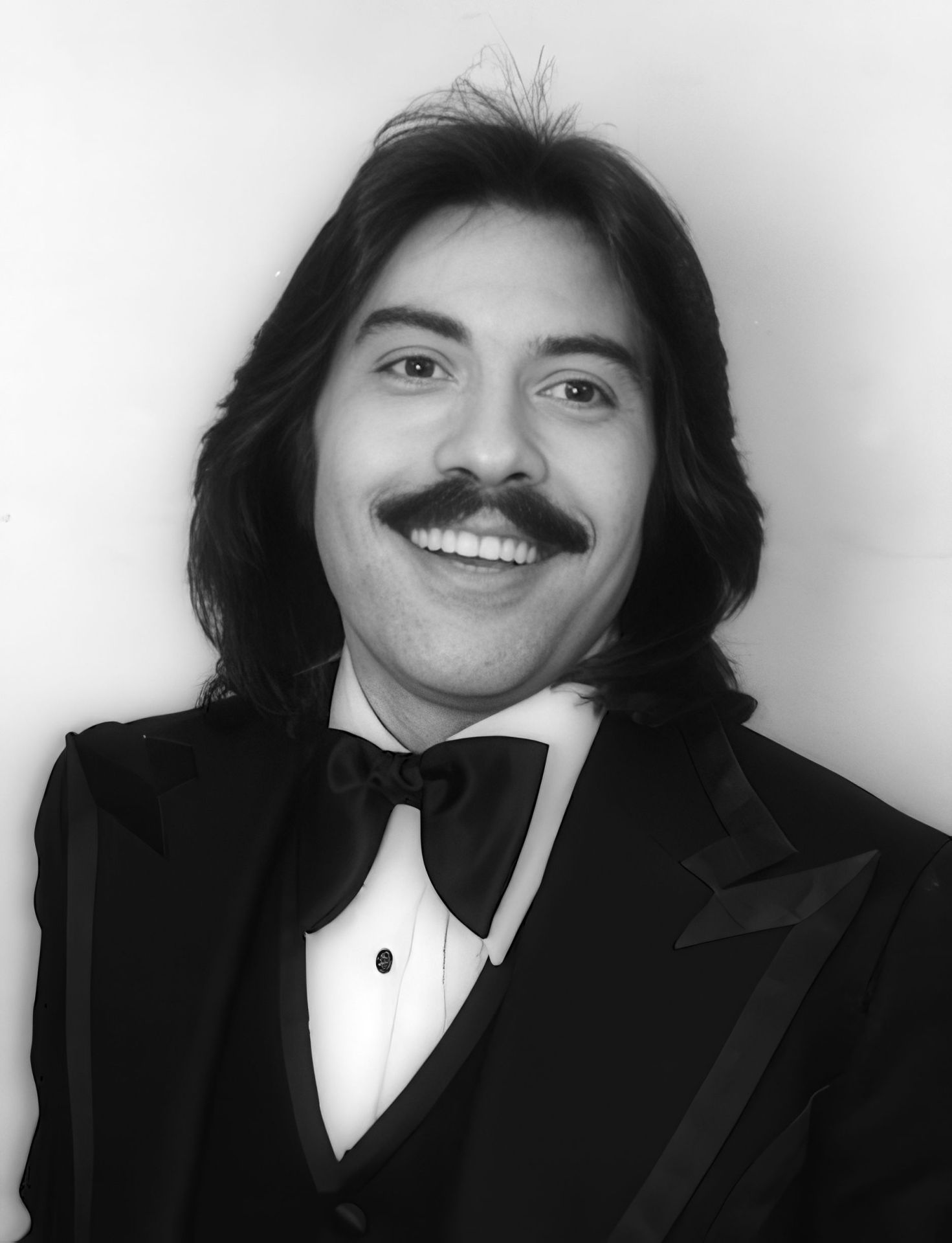 Black and white portrait of a smiling young man wearing a tuxedo and bow tie, featuring long, dark hair and a mustache.