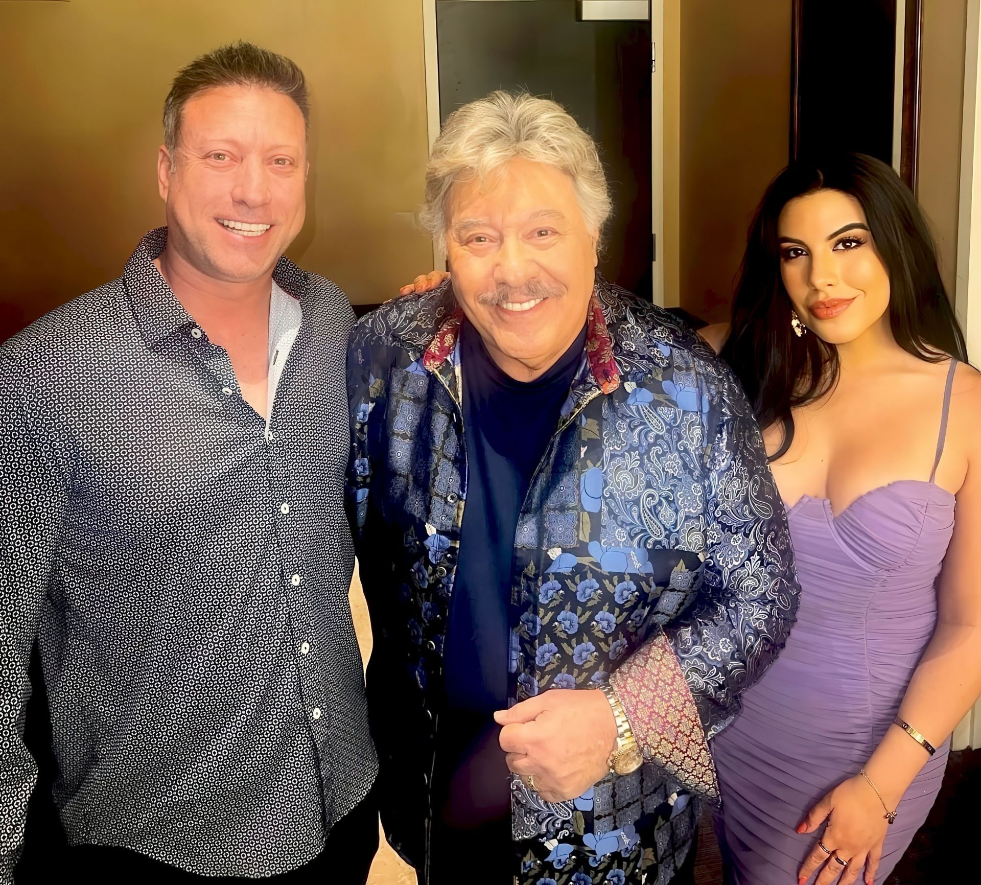 Three people posing together in a well-lit room. On the left, a man with dark hair and a patterned shirt is smiling. In the center, an older man with gray hair and a colorful patterned blazer is grinning and holding his hand out. On the right, a woman with long dark hair is wearing a form-fitting purple dress and smiling at the camera.