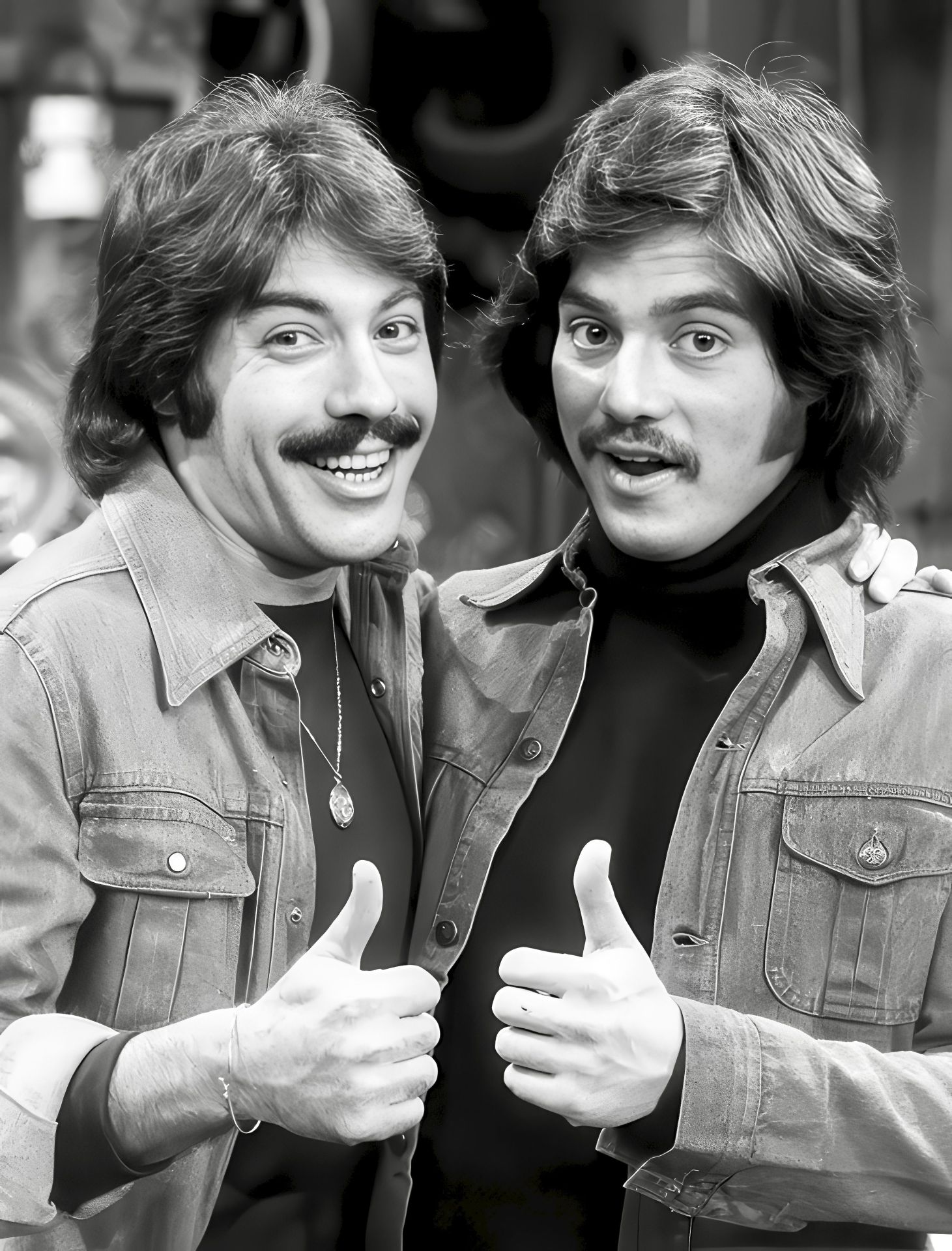 Black and white photo of two young men with mustaches, smiling and giving thumbs up while wearing matching denim jackets.