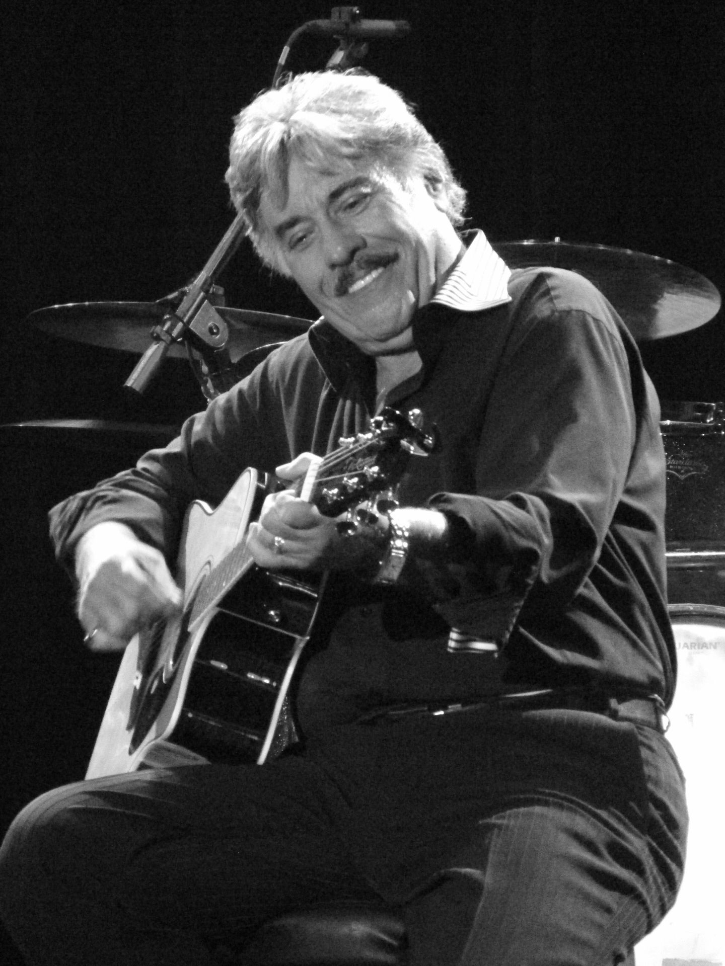Black and white image of a smiling male musician playing an acoustic guitar on stage, with a microphone and drums in the background.