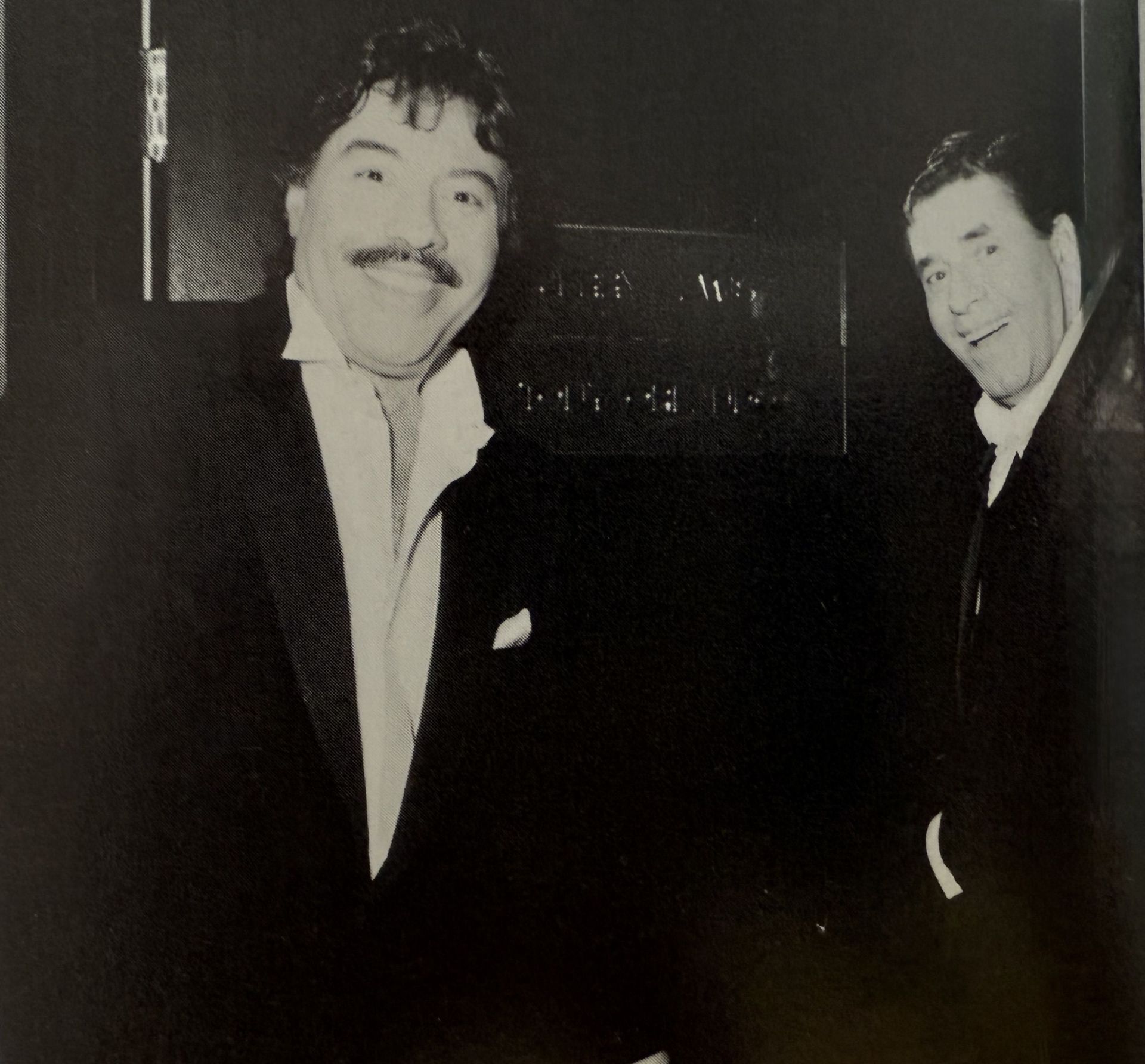 Black and white photo of two smiling men in tuxedos, standing side by side, with a sign in the background.