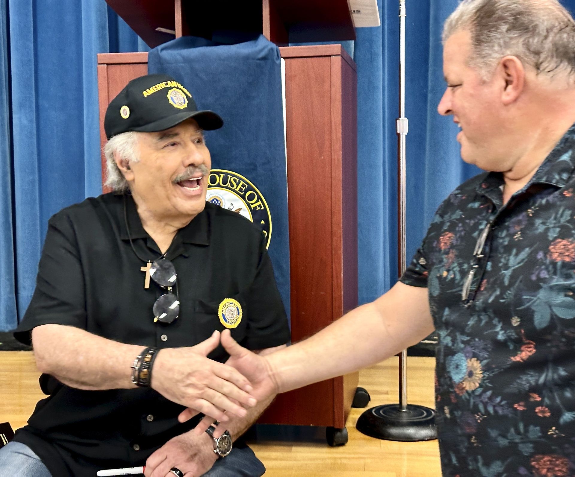 A smiling man with gray hair and glasses shakes hands with another man in a floral shirt during a heartfelt interaction. They are seated in front of a podium with a seal and a blue curtain backdrop.