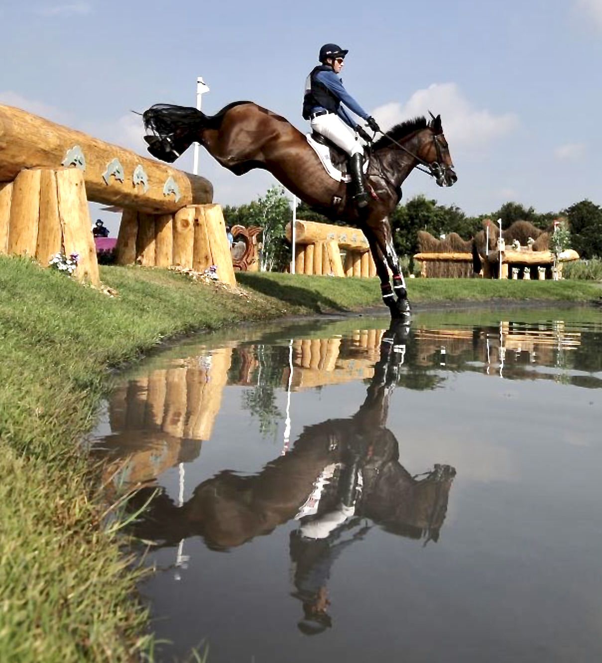 A horse and rider jumping over a log obstacle near a body of water, with the rider's reflection visible in the water.