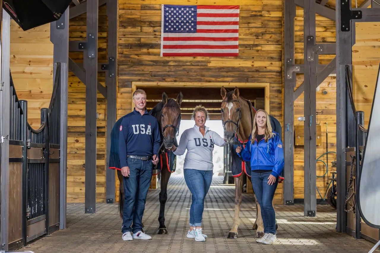 Three equestrians pose inside a wooden barn holding their horses, with an American flag displayed above them.