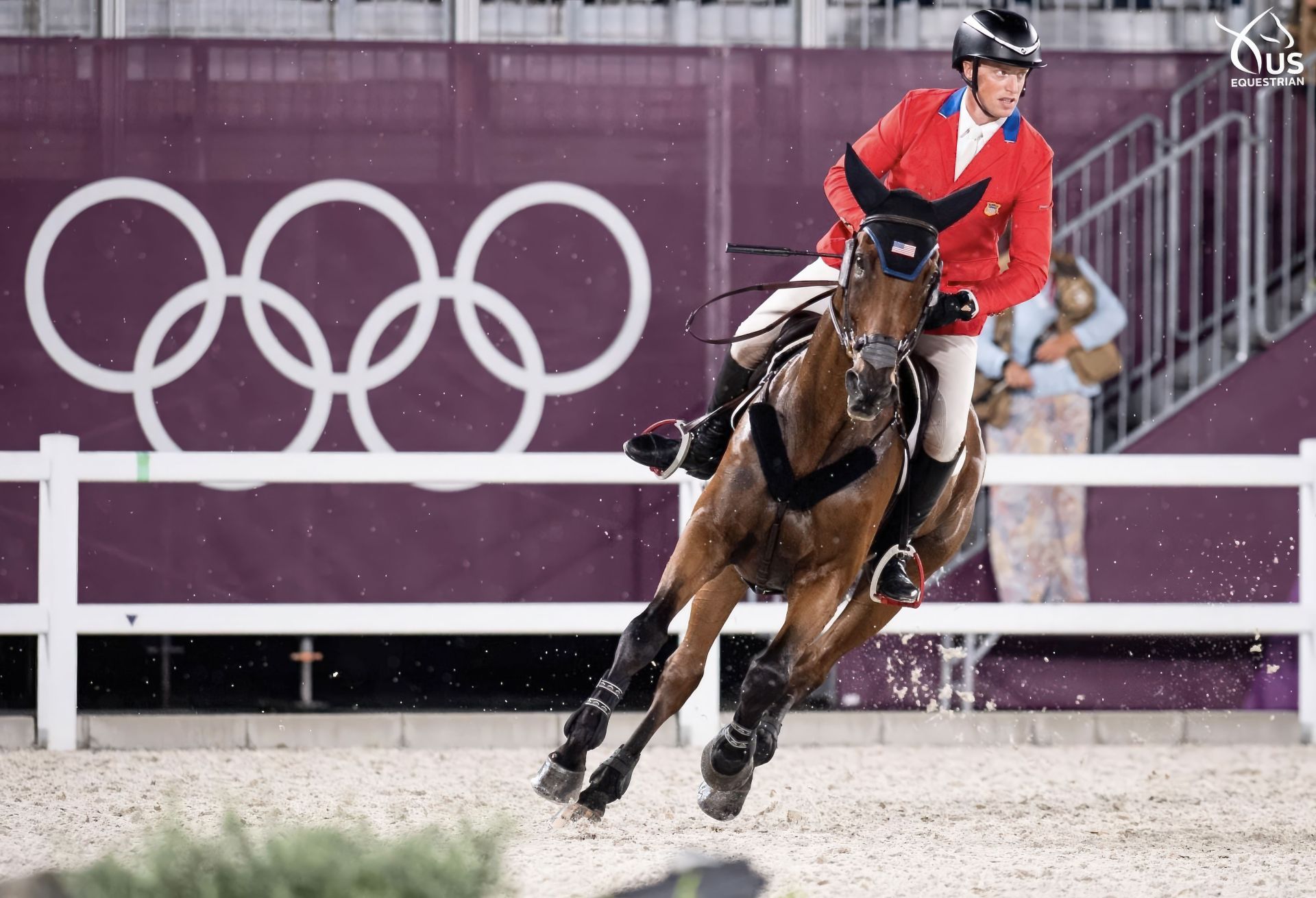 A rider in a red jacket competes on a horse during an equestrian event at the Olympics, with the Olympic rings in the background.