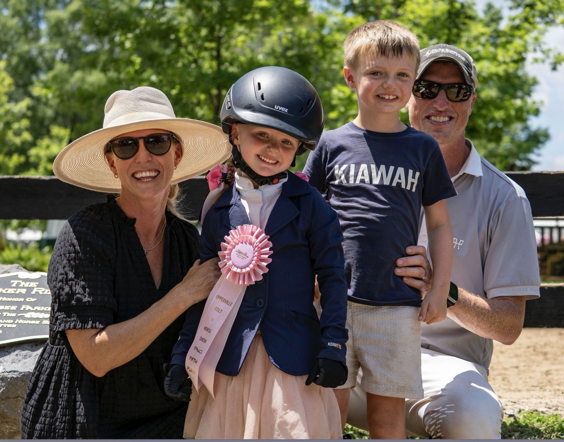 A family portrait featuring a woman wearing sunglasses and a wide-brimmed hat, a young girl in riding attire holding a pink ribbon, a young boy in a blue shirt, and a man in a polo shirt, all smiling and posing outdoors near a wooden fence surrounded by greenery.