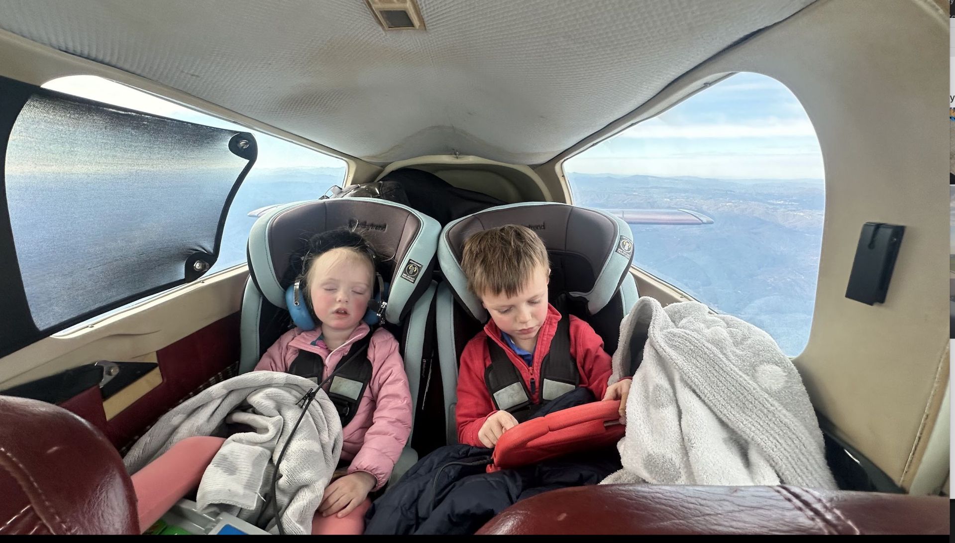 Two young children seated in an aircraft, one sleeping and the other focused on a tablet, with scenic views visible through the window.