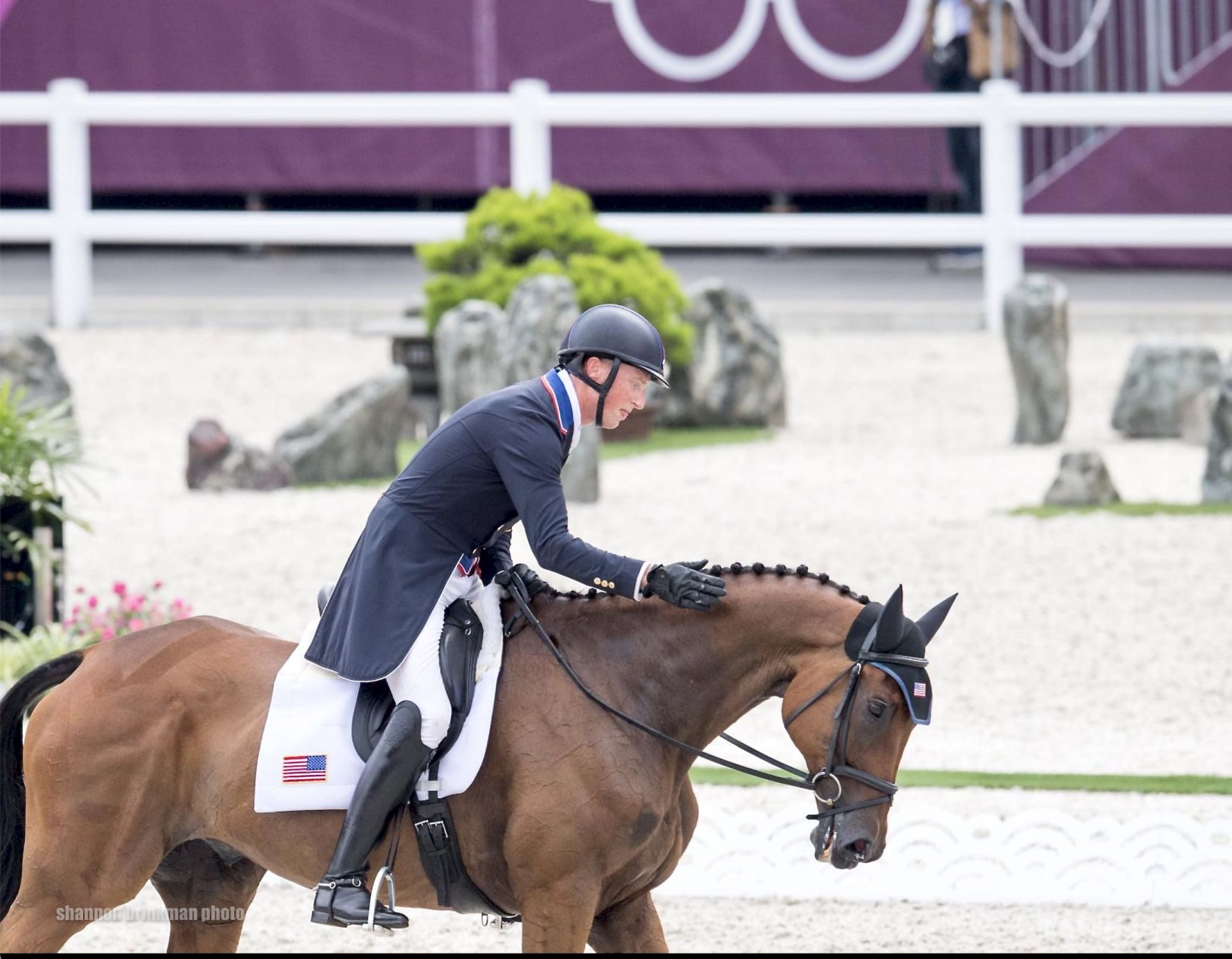 A male equestrian in a formal riding outfit gently pats the neck of his horse, displaying a moment of connection during a competition, with a blurred background of stone decorations and an audience area.