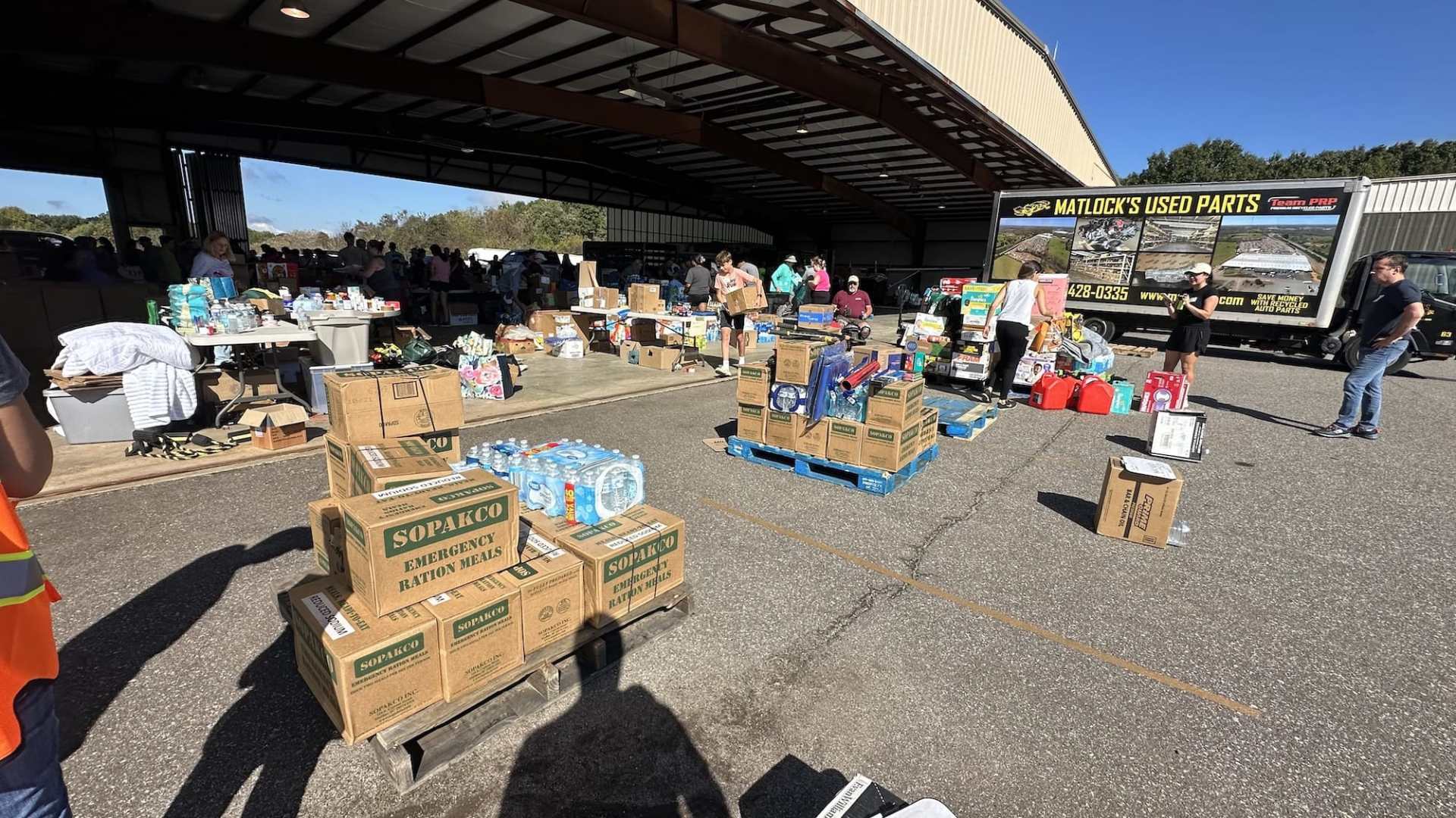 A group of people organizing supplies for disaster relief outside a warehouse, with stacks of boxes containing food, water, and other essential items.
