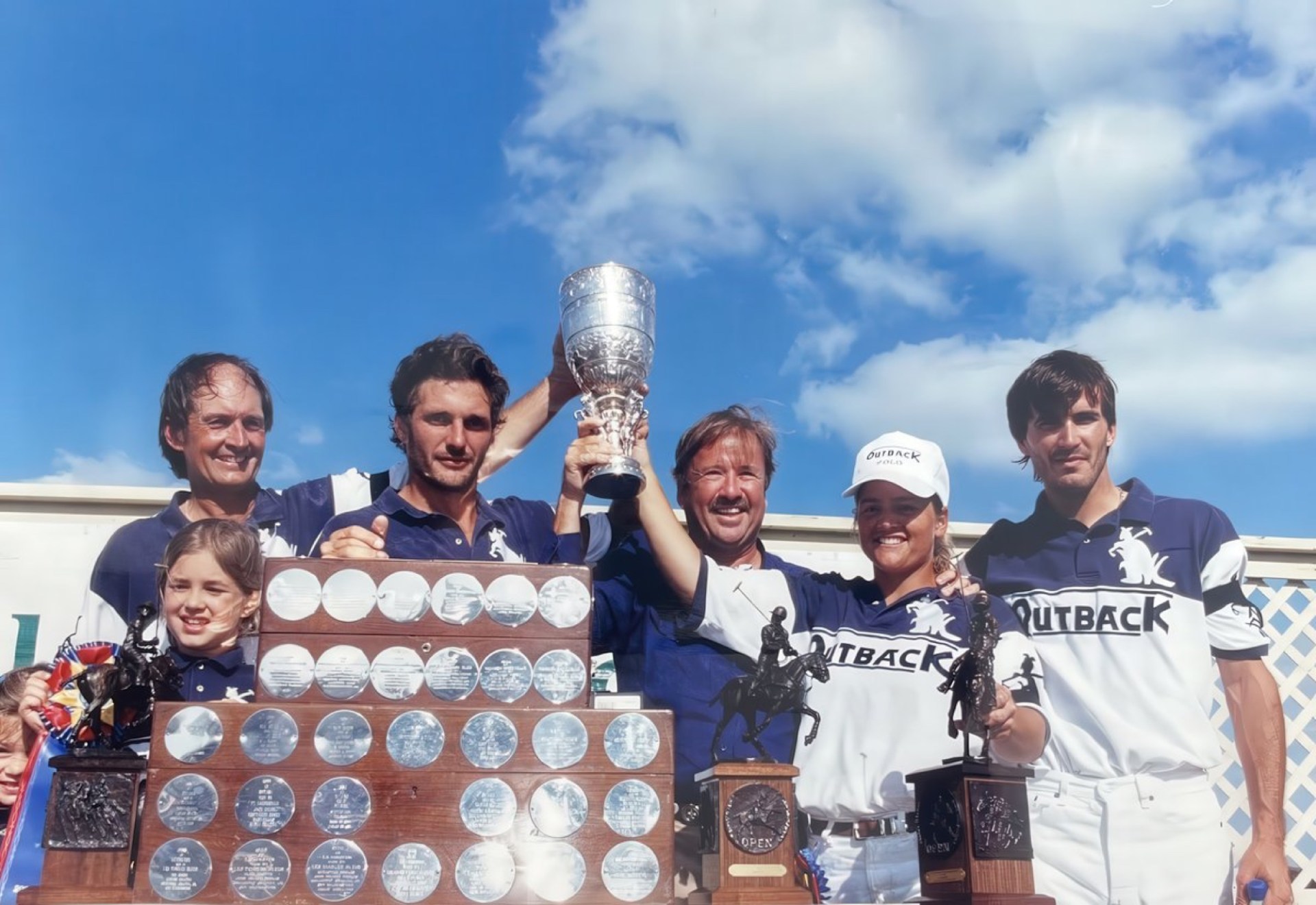 A group of six people celebrating with trophies and a large plaque covered in engraved medals, under a blue sky with scattered clouds.
