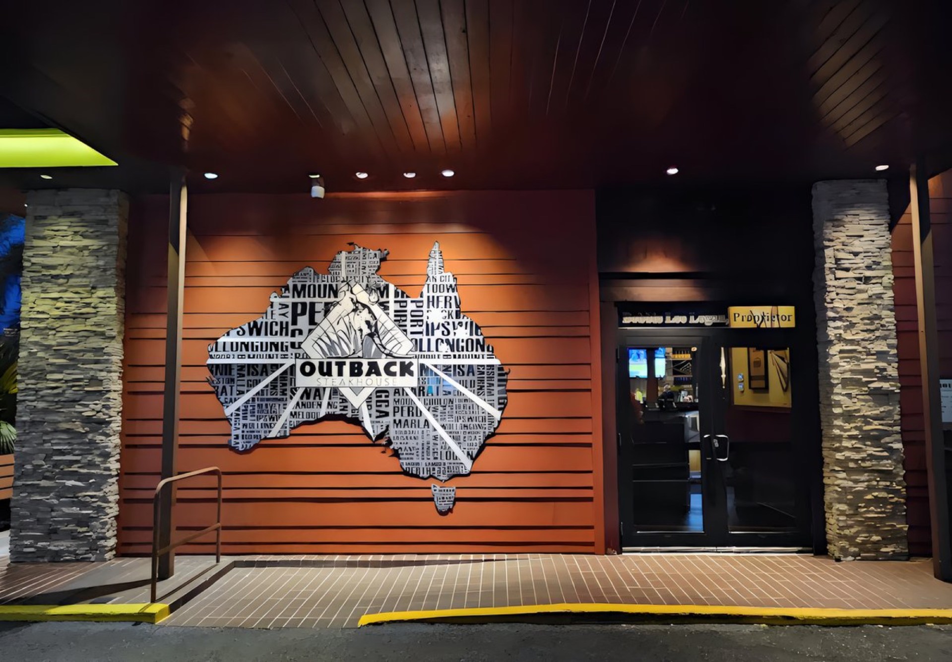 Exterior view of an Outback Steakhouse restaurant featuring a large decorative map of Australia made from text elements, with stone accents on the walls.
