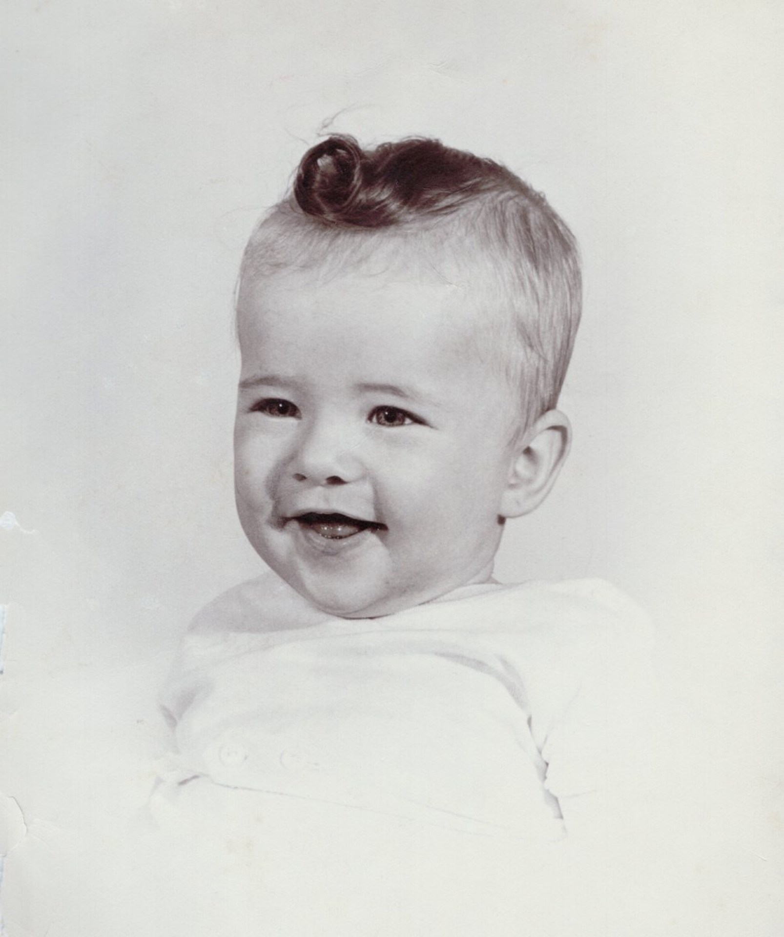 A smiling baby with light hair and a small curl on top, wearing a white outfit, against a plain background.