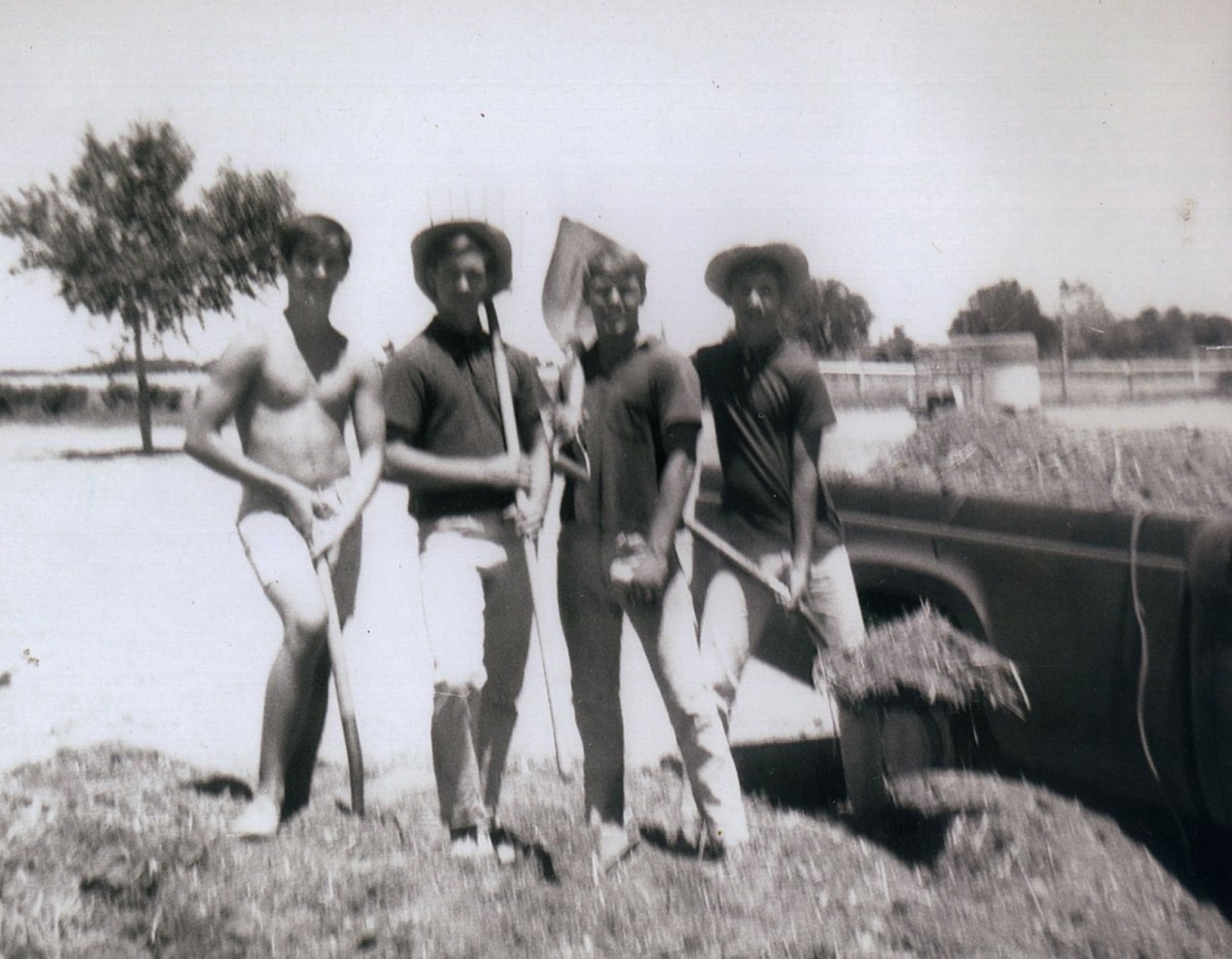 Four young men standing outdoors on a farm, two holding tools and one shirtless, with a truck and trees in the background.
