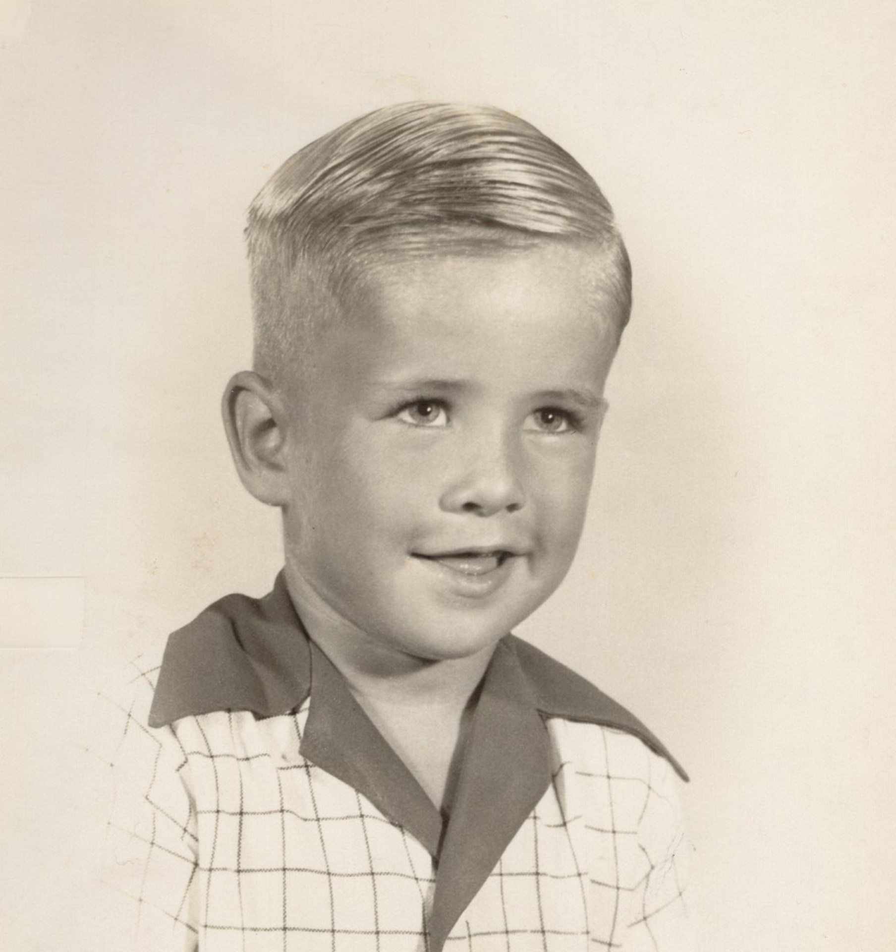 A smiling young boy with short, slicked-back hair, wearing a checkered shirt with a colored collar, against a light background.