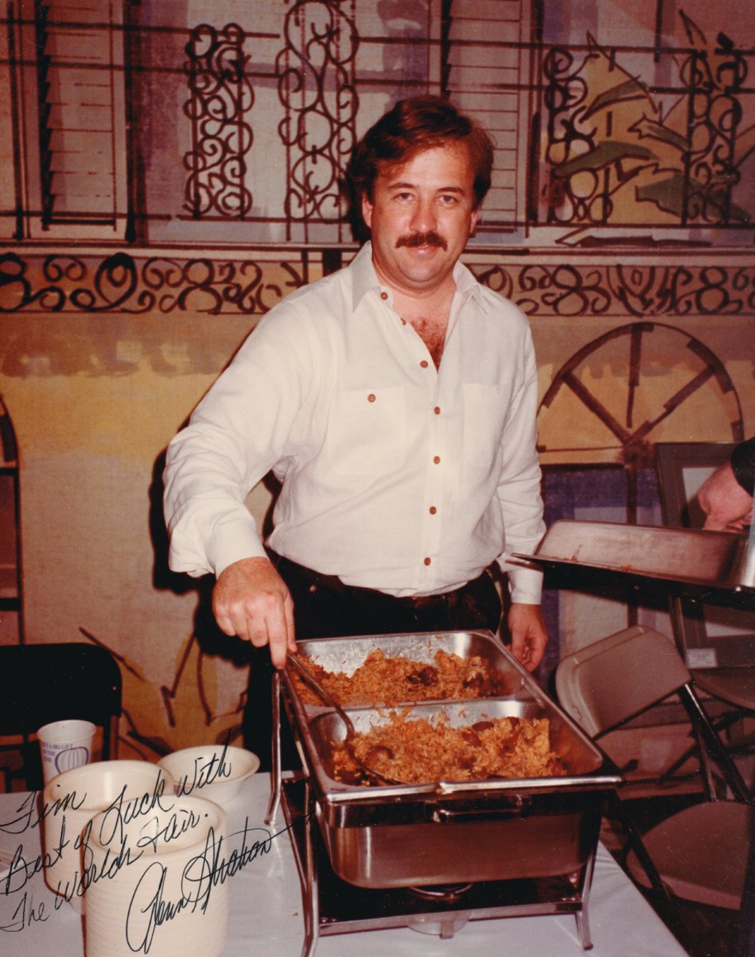 A man with a mustache wearing a white shirt is serving food from a chafing dish at an event, with decorative background elements visible.