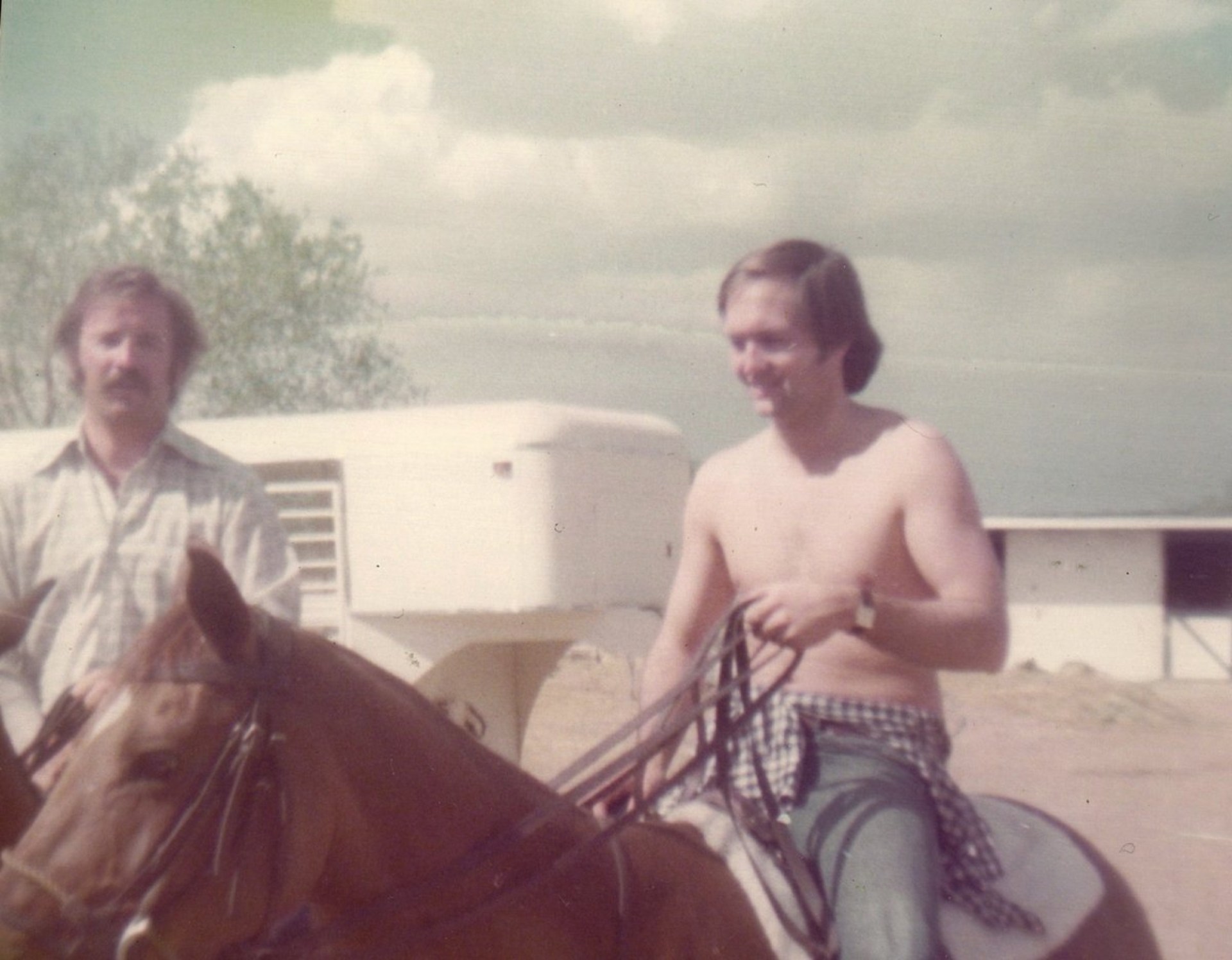 Two men outdoors near a horse. One man is shirtless and riding the horse, while the other man stands beside him, looking at the camera.