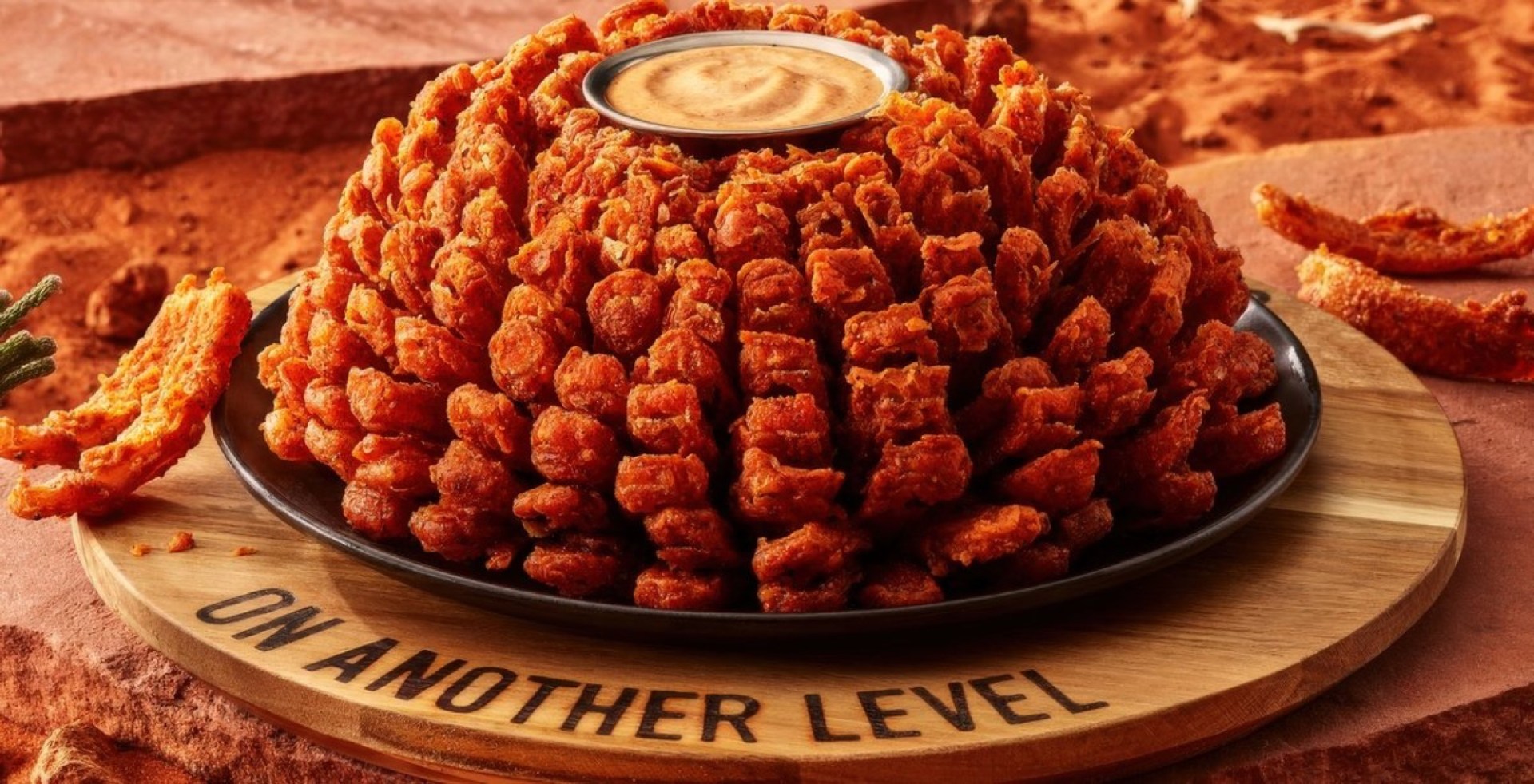 A large plate of crispy, fried chicken pieces arranged in a mound, served with a small bowl of dipping sauce on top, presented on a wooden platter with the words 'ON ANOTHER LEVEL' engraved.