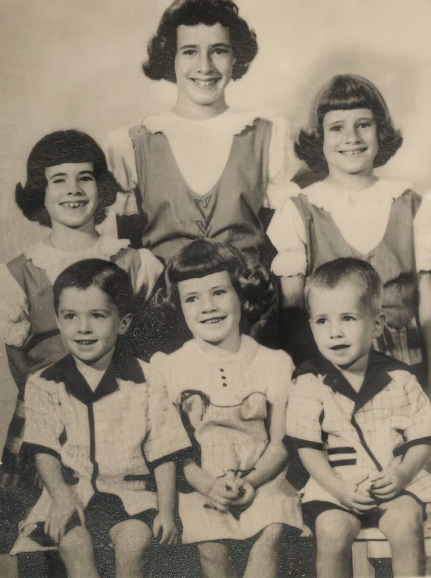 A black and white photograph of six children, three boys and three girls, posed together. The older girls are standing in the back row, while the younger children are seated in front, all smiling and dressed in vintage clothing.