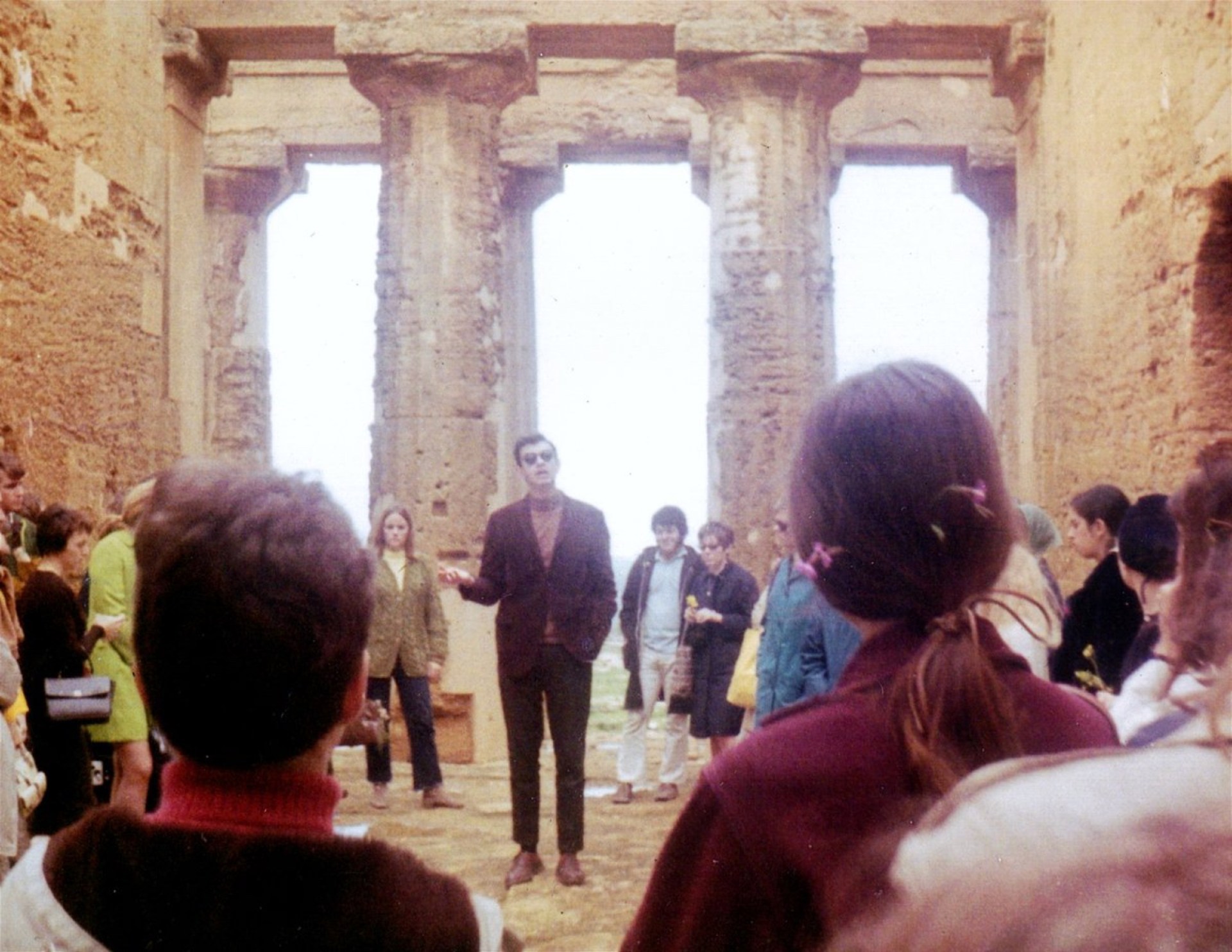 A group of people gathered inside a historical site, with a man in the foreground addressing the crowd. The setting features ancient stone columns and walls, suggesting an archaeological or cultural significance.