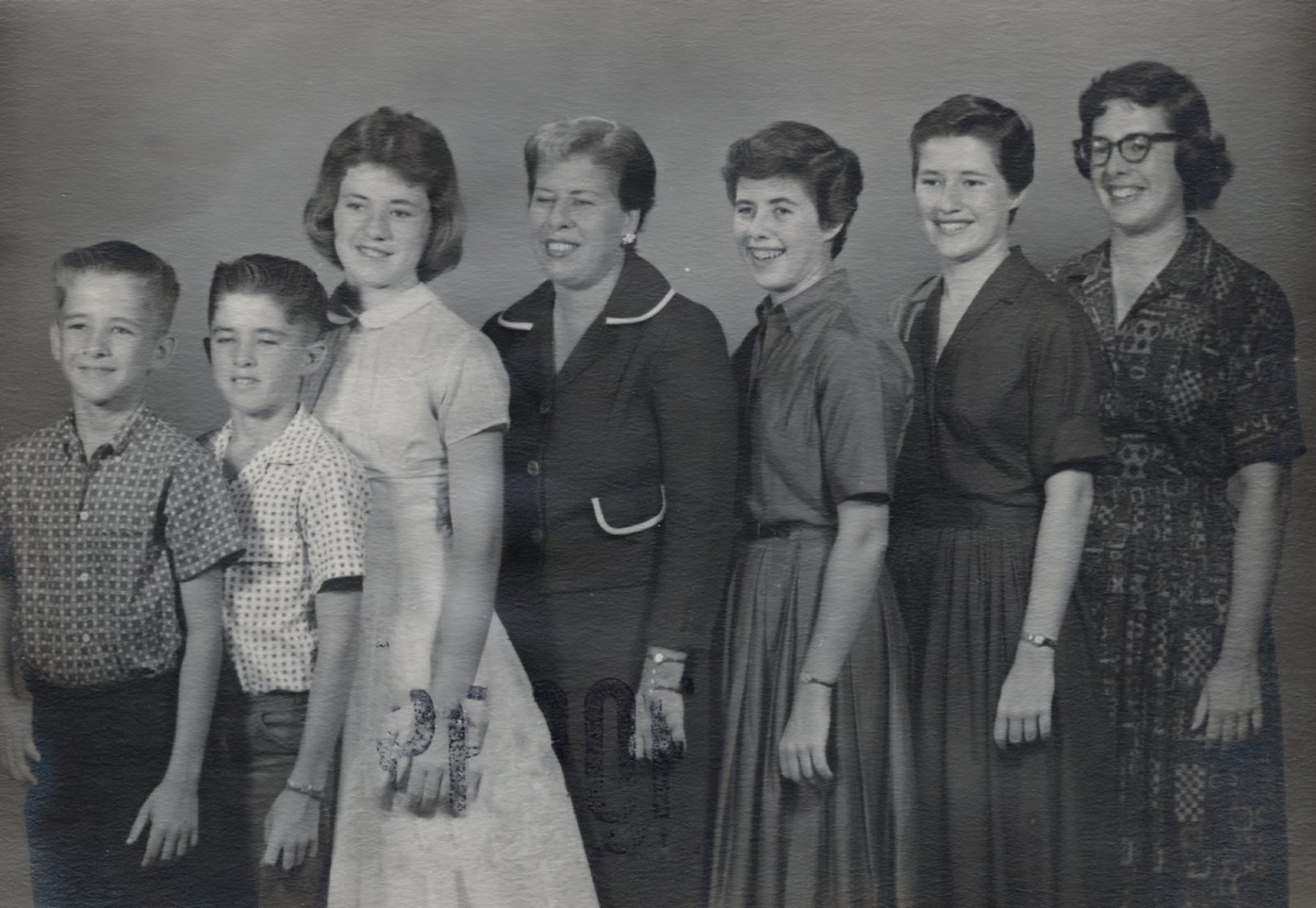 A black and white photo of a group of six children and one adult woman standing in a line, smiling. The children are wearing casual clothes, while the woman is dressed in a dark blouse with a white trim.