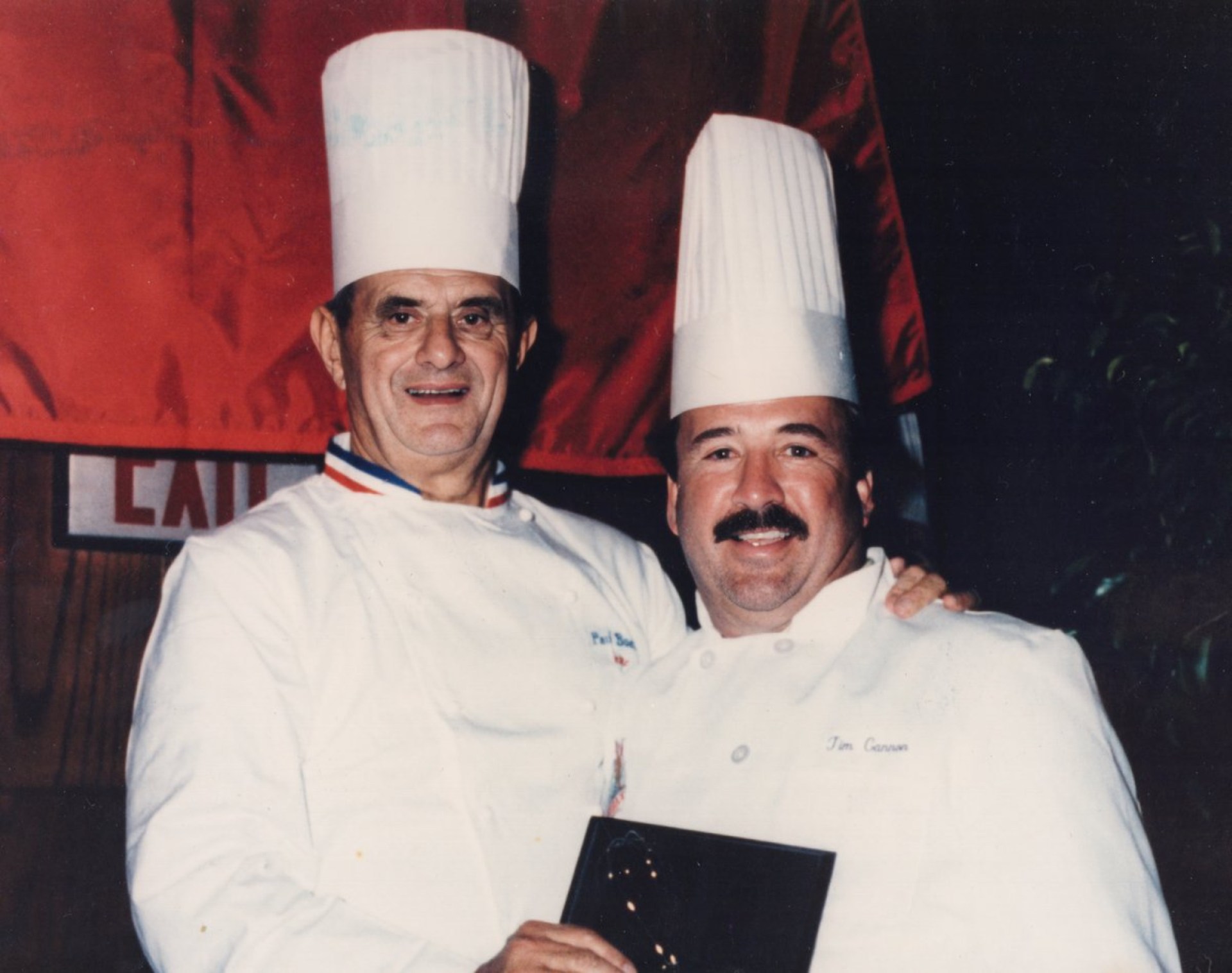 Two chefs smiling together, one wearing a chef's hat and white coat, holding an award, with a red backdrop.