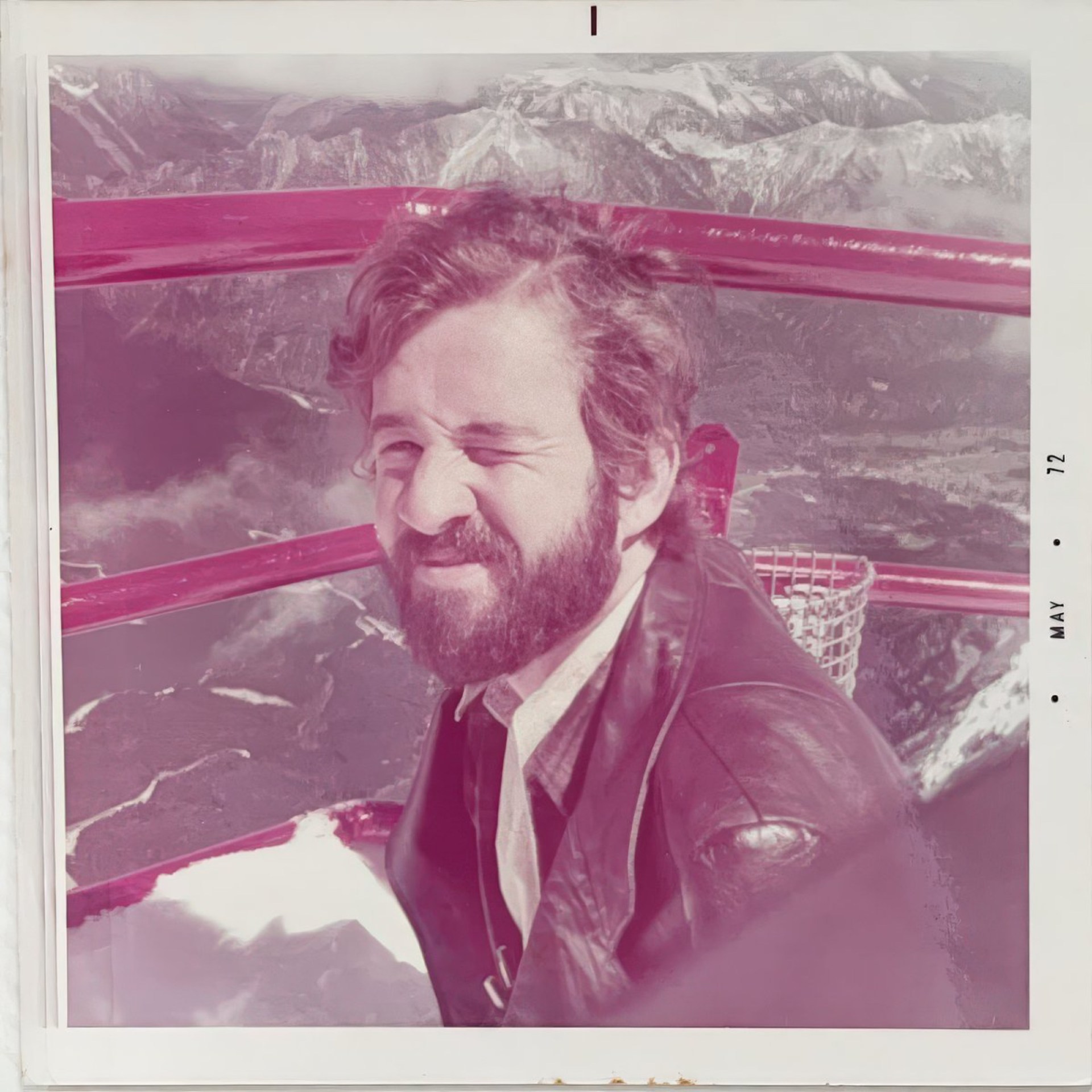 A man with a beard smiles and winks while sitting in a gondola with scenic mountainous views in the background.