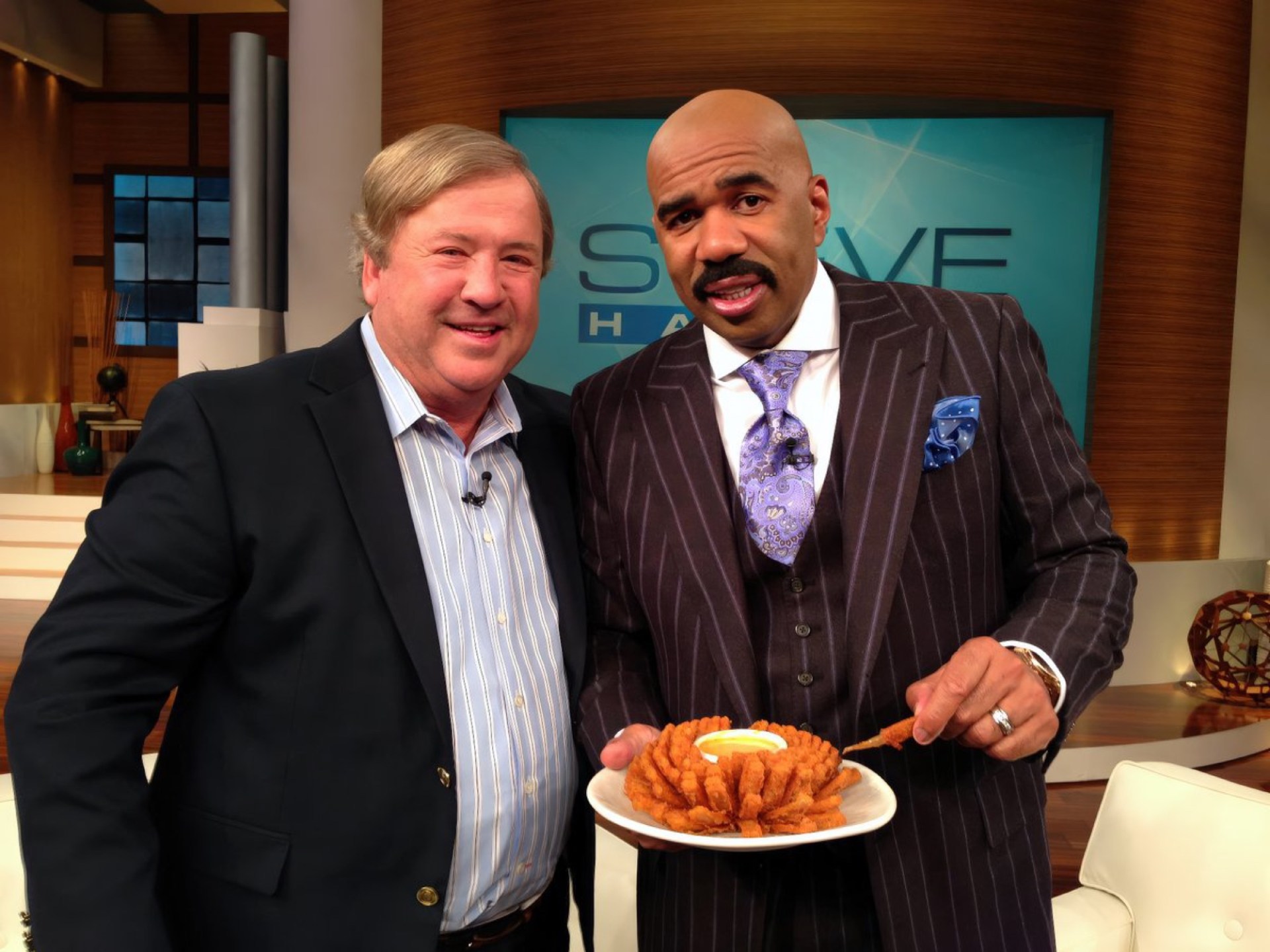 Two men smiling together in a studio setting, one holding a plate of fried food.
