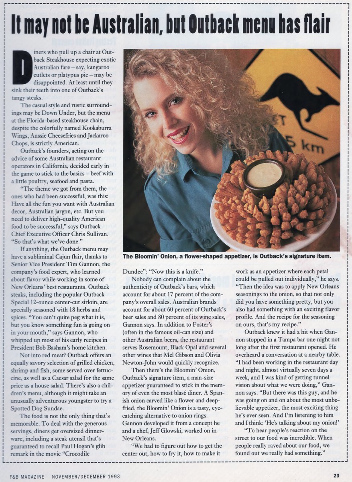 A smiling woman with curly hair holds a large platter of Bloomin' Onion, a flower-shaped appetizer from Outback Steakhouse, in front of an Australian-themed sign.
