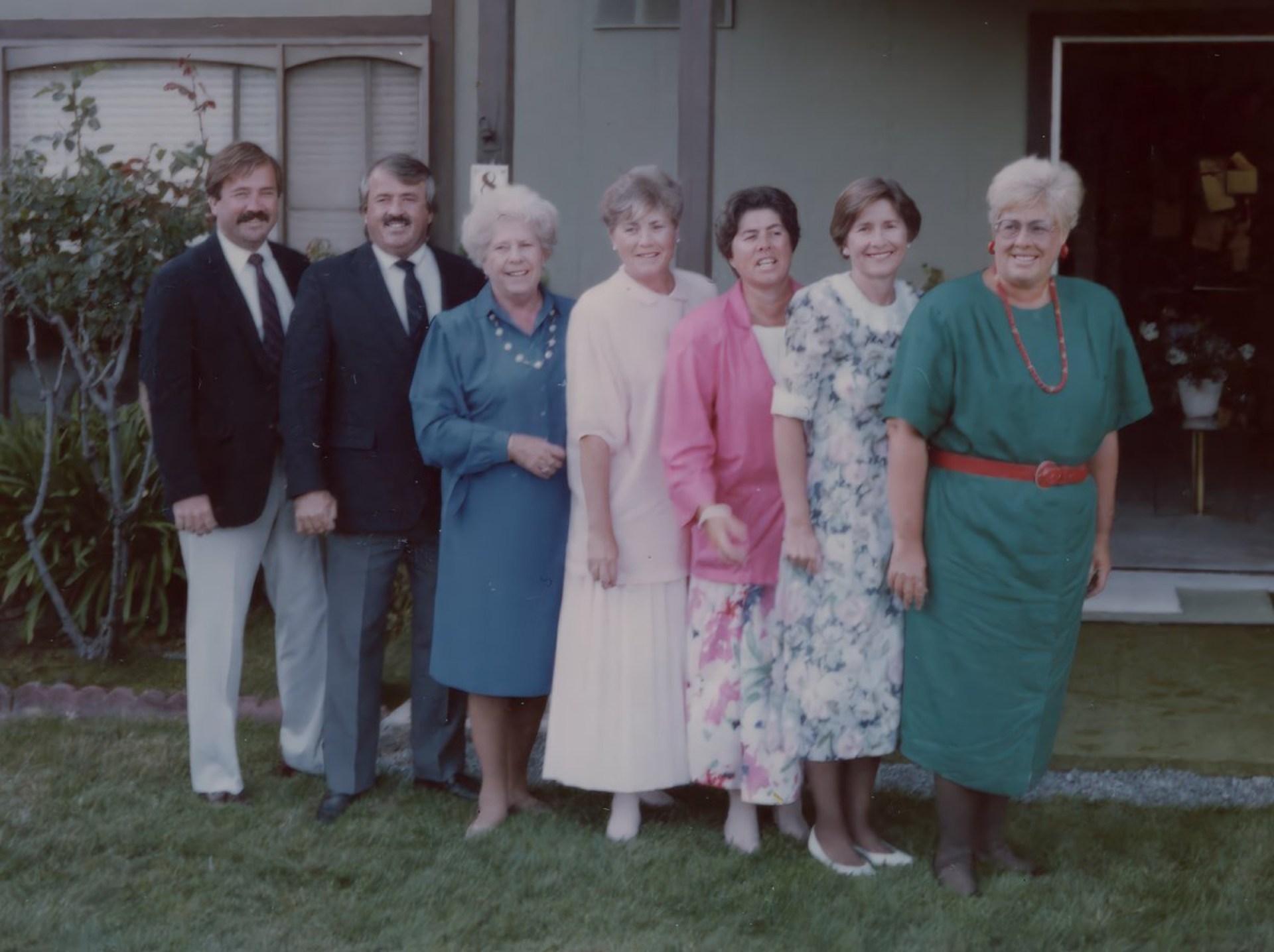 A group of seven people standing side by side outdoors in front of a house, smiling. They are wearing a variety of colorful outfits, including suits and dresses, with some casual styles. The setting features a green lawn and shrubbery in the background.