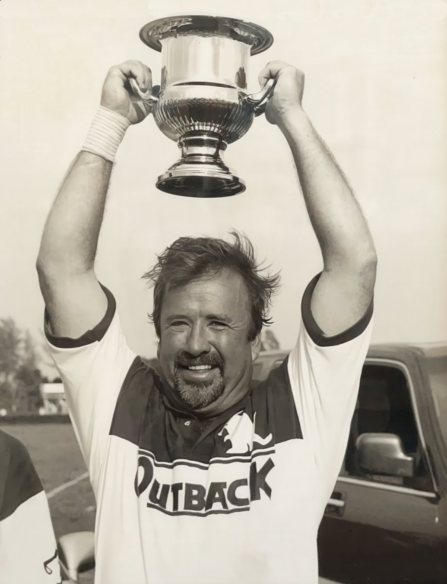 A man wearing a polo shirt with 'Outback' logo is joyfully holding a trophy above his head, smiling widely in an outdoor setting.