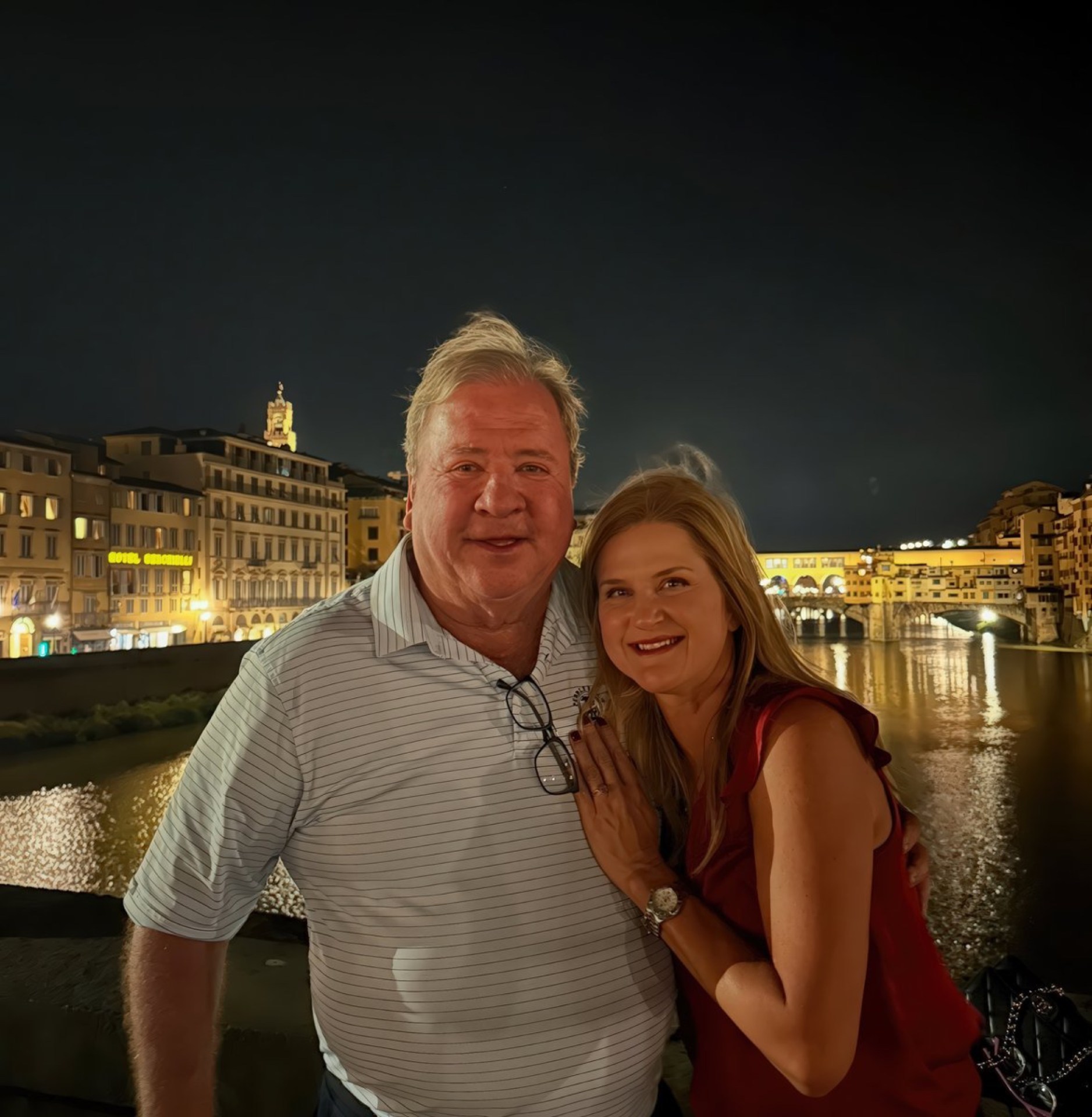 A man and woman smiling together at night, with the illuminated Ponte Vecchio bridge and city buildings in the background.