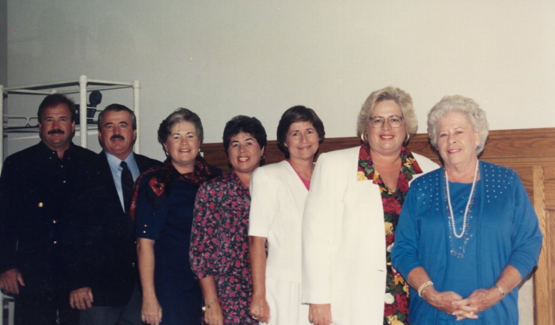 A group of seven people standing together, smiling in a well-lit indoor setting. They are dressed in various colorful outfits, with three men and four women. The background is simple and features a wooden panel.