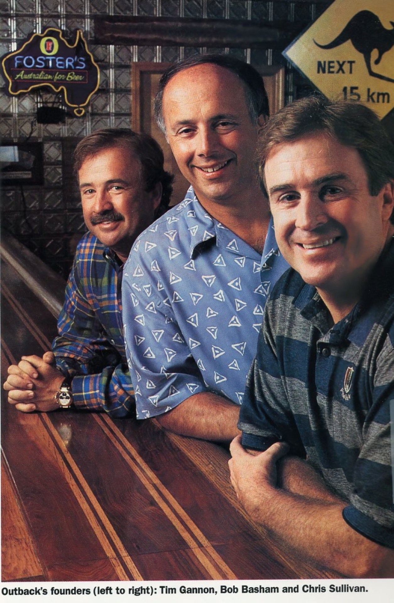 Three men posing together at a wooden bar with a Foster's beer sign and a kangaroo crossing sign in the background.