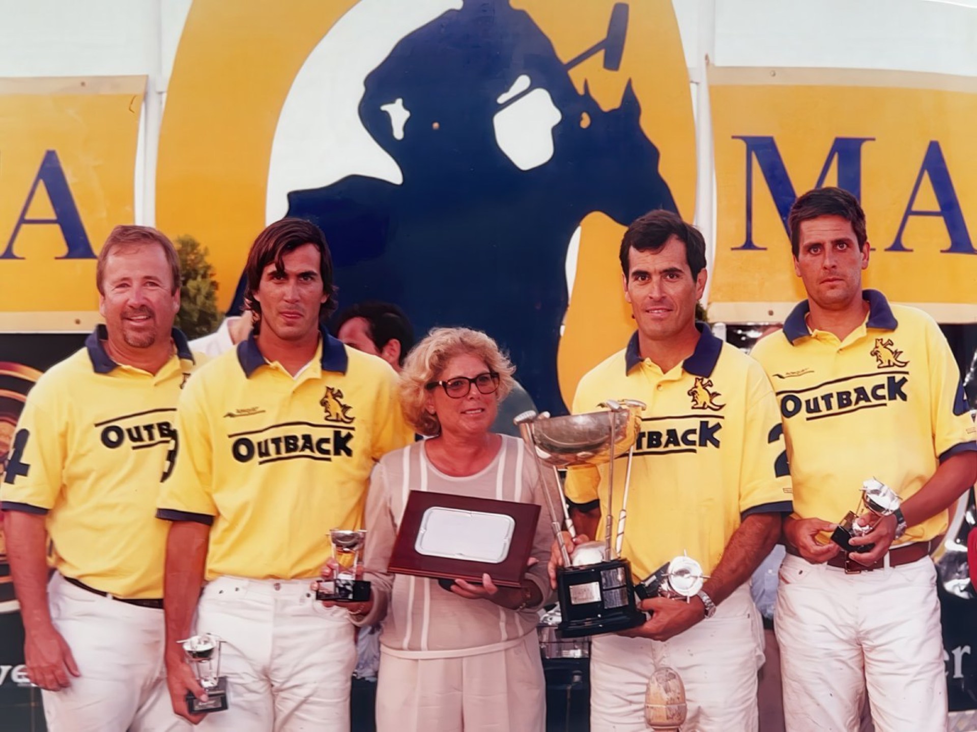 Group photo of five individuals, including four men wearing yellow polo shirts with 'Outback' branding, standing with a woman holding multiple awards and trophies, set against a promotional backdrop.