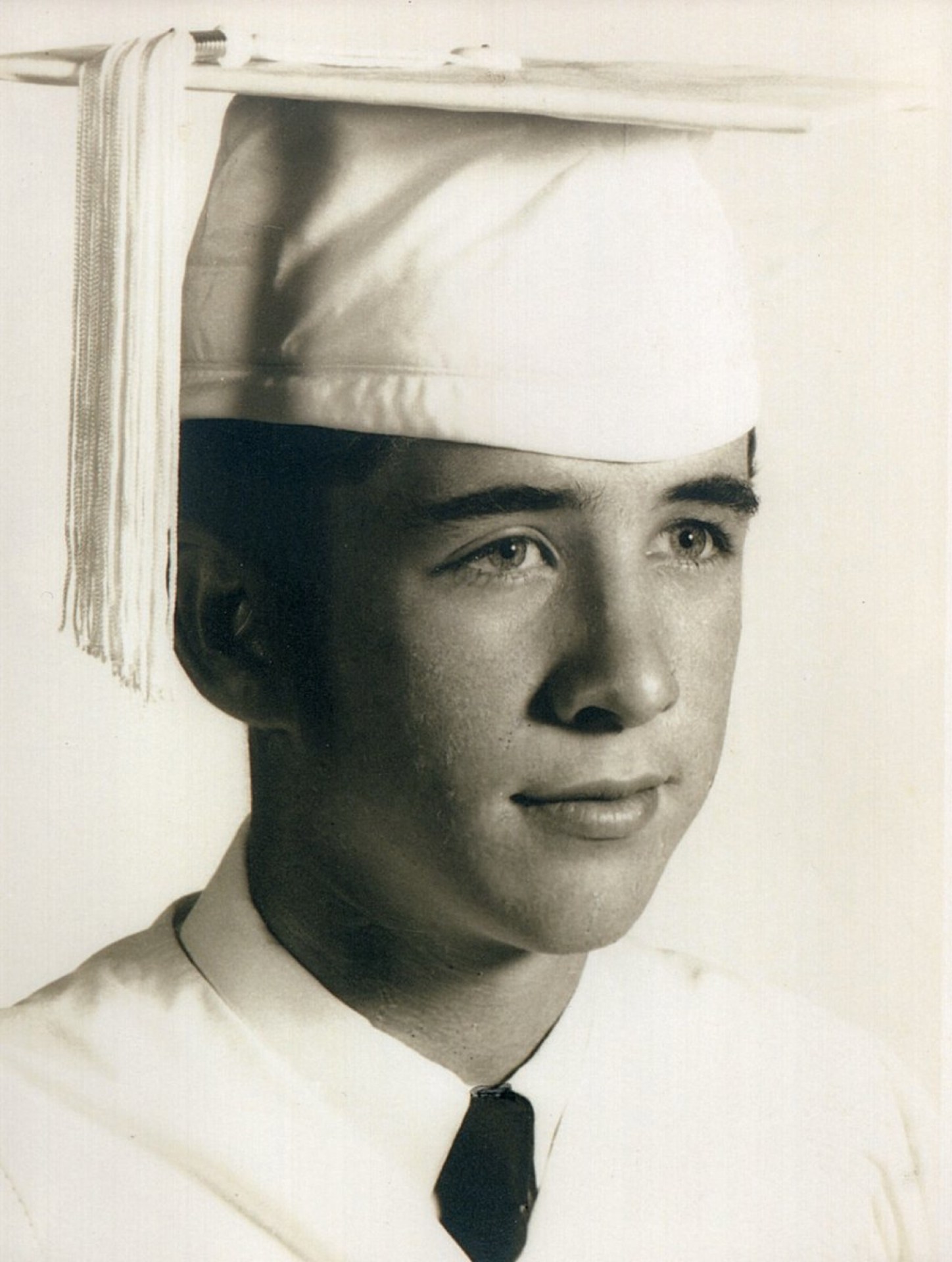 A young male graduate in a white cap and gown, posing for a portrait with a serious expression.