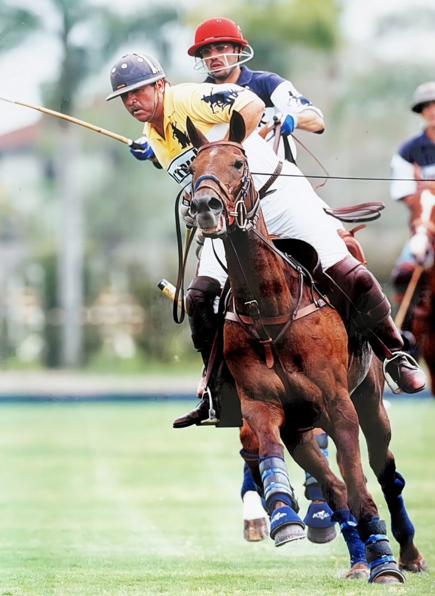 A polo player in a yellow shirt riding a brown horse, actively engaging in a game while swinging a polo stick. Another player in a red helmet is closely following behind.