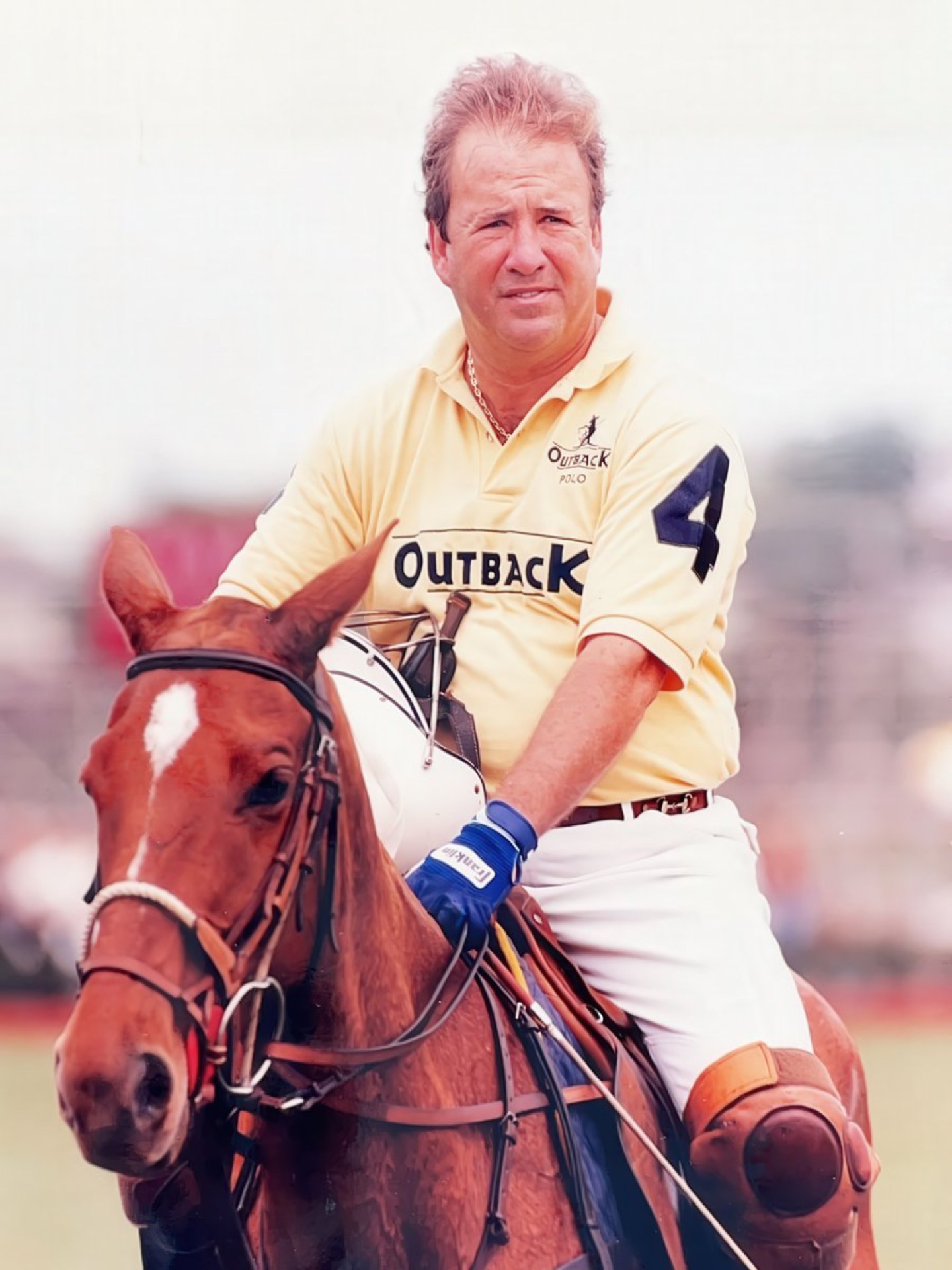 A man wearing a yellow polo shirt with the word 'Outback' and the number 4 rides a brown horse, holding a polo mallet, in a polo match setting.