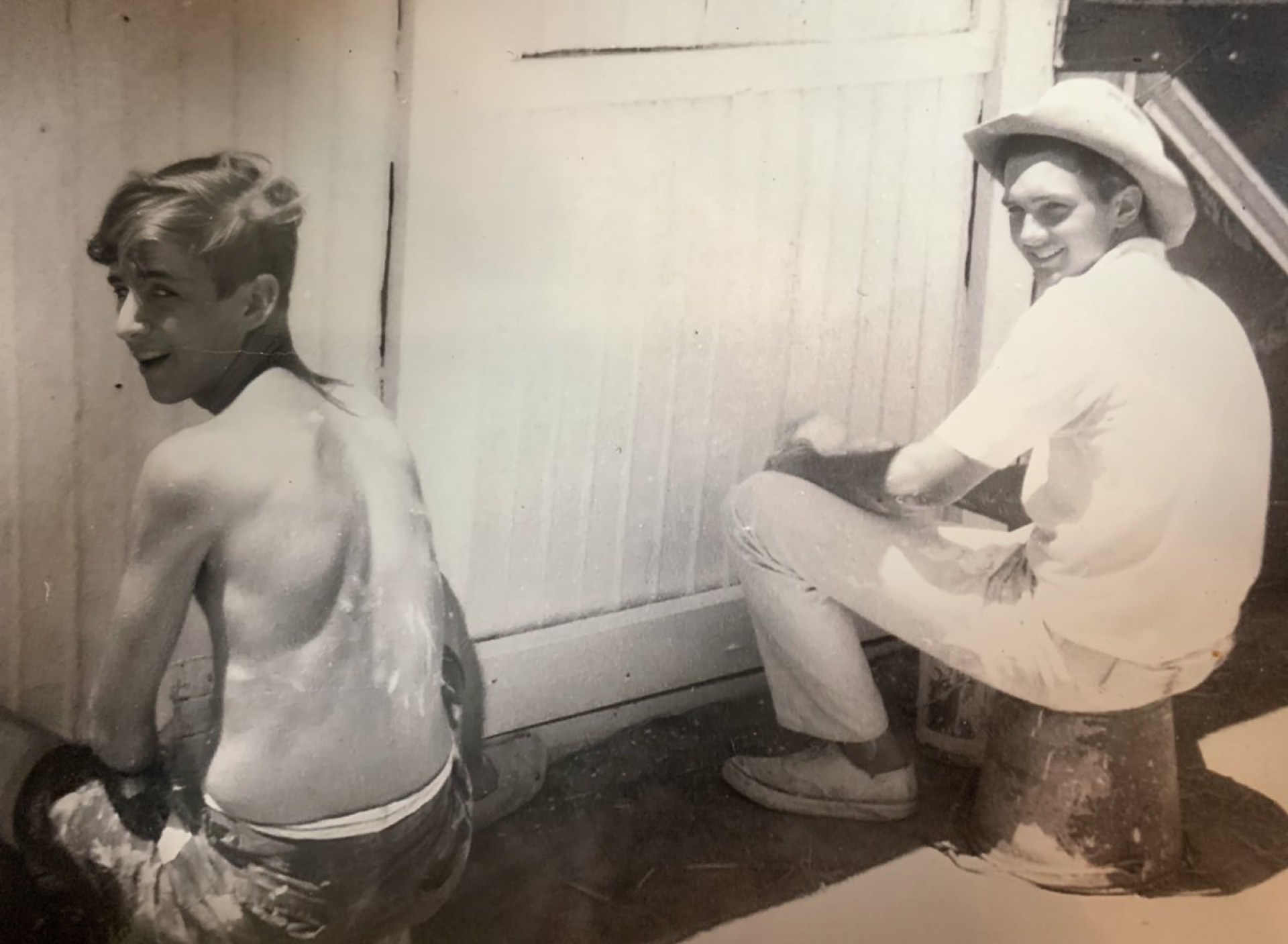 Two young men seated outside near a building, one with a bare back looking over his shoulder and the other wearing a white shirt and cowboy hat, both engaged in a casual activity.