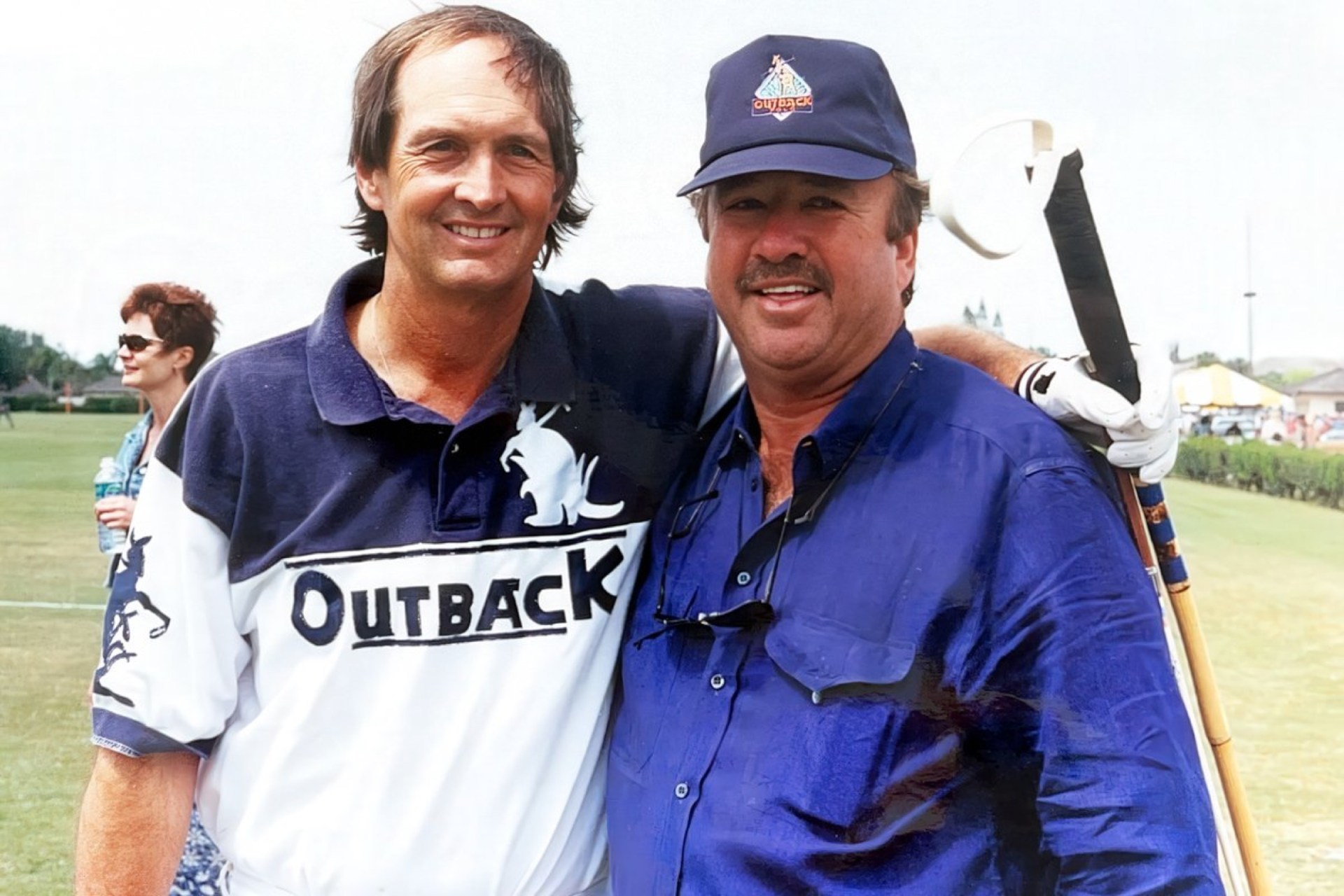 Two men smiling together, one wearing a black and white Outback polo shirt, the other in a blue shirt and cap. A woman in sunglasses stands in the background.