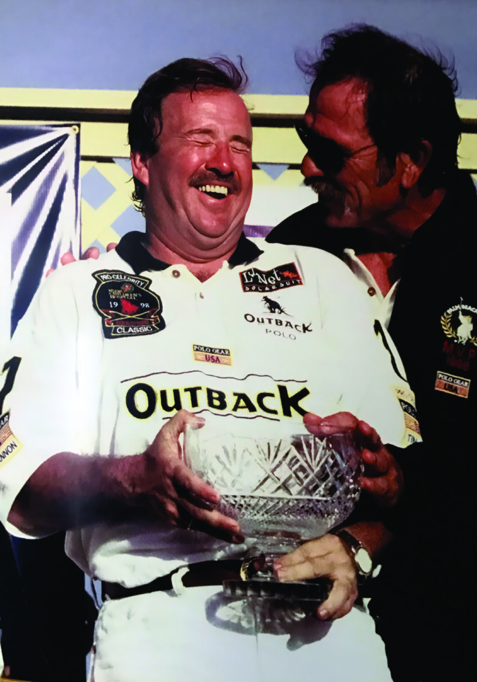 Two men celebrating on a podium, one laughing and holding a trophy, while the other leans in close, sharing a joyful moment.